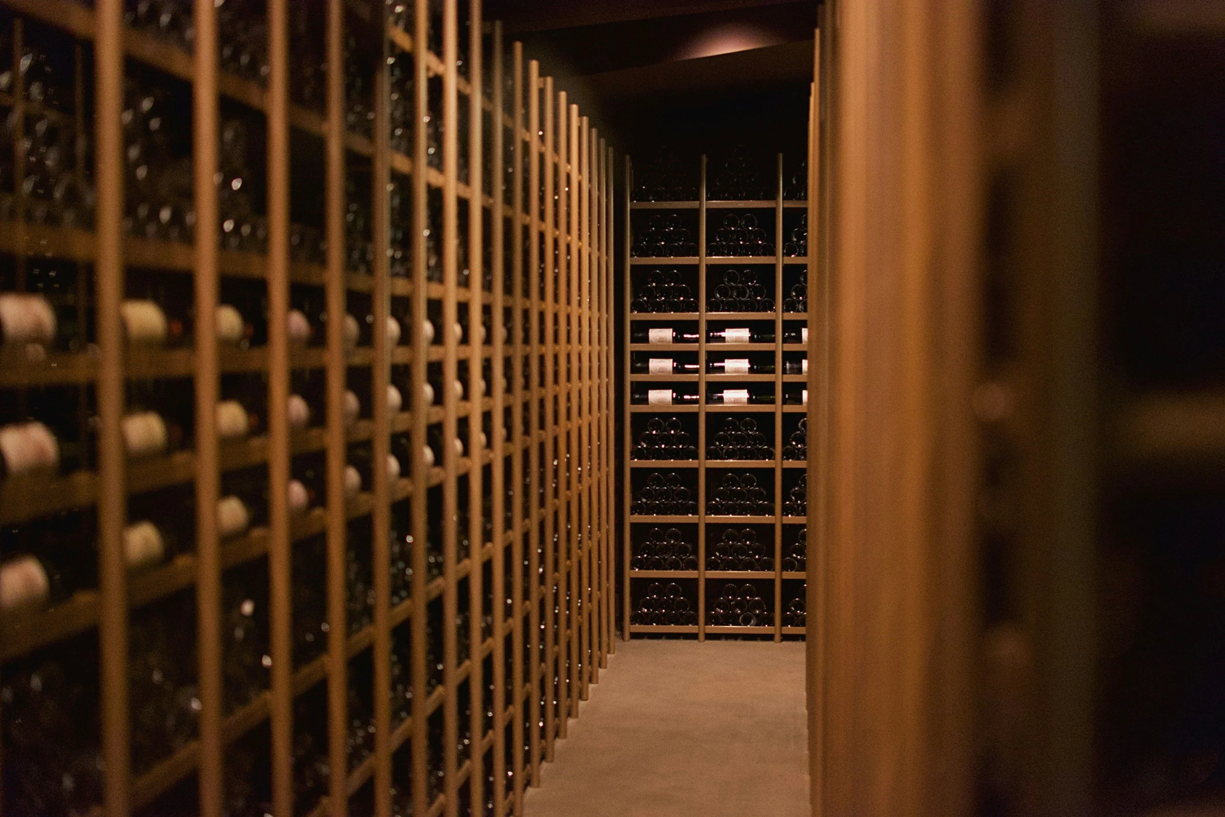 A wine cellar with wooden racks filled with wine bottles, viewed through a narrow aisle.