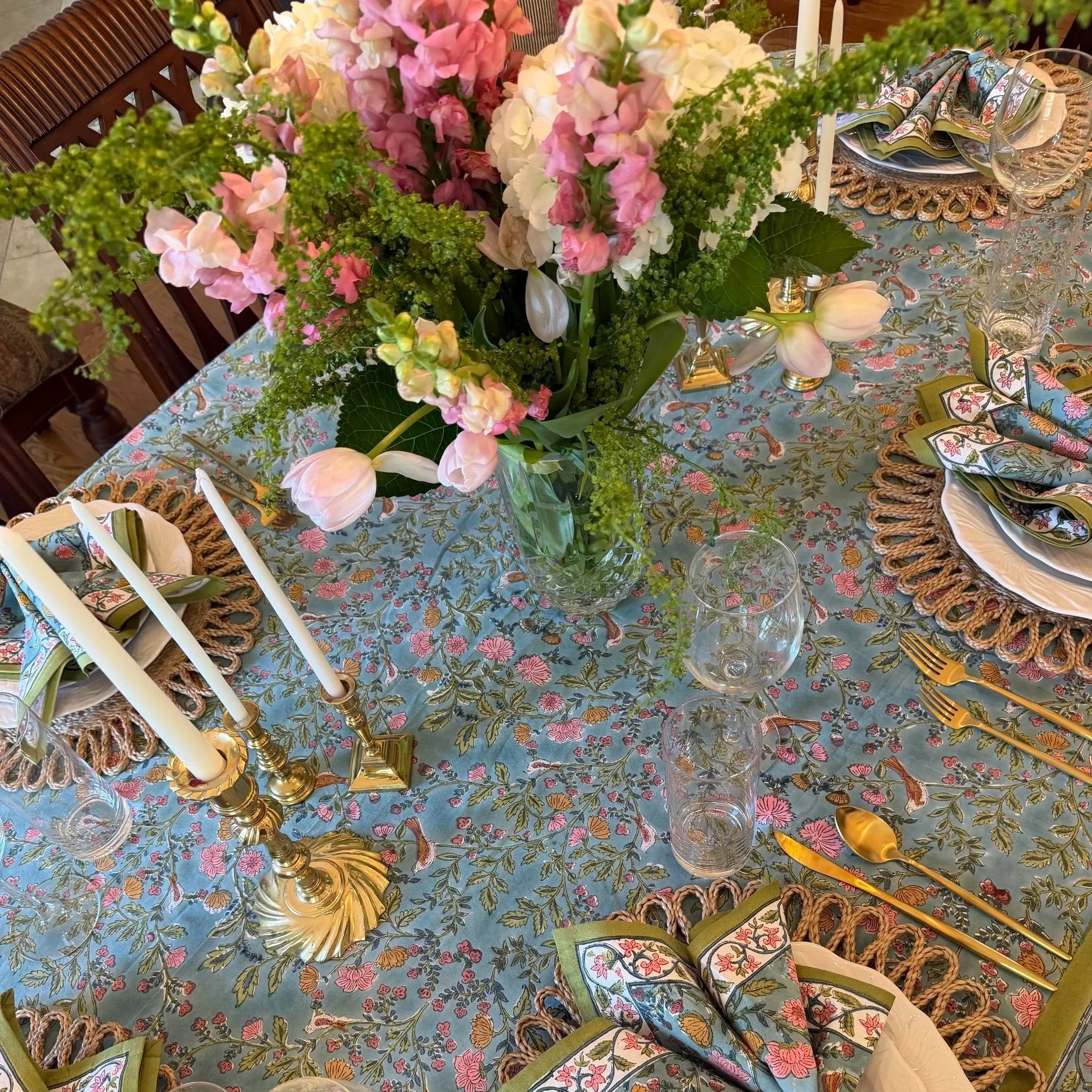 A dining table decorated with a floral centerpiece featuring pink, white, and green flowers, surrounded by place settings with patterned napkins, glassware, and gold flatware on a tablecloth with a floral pattern.