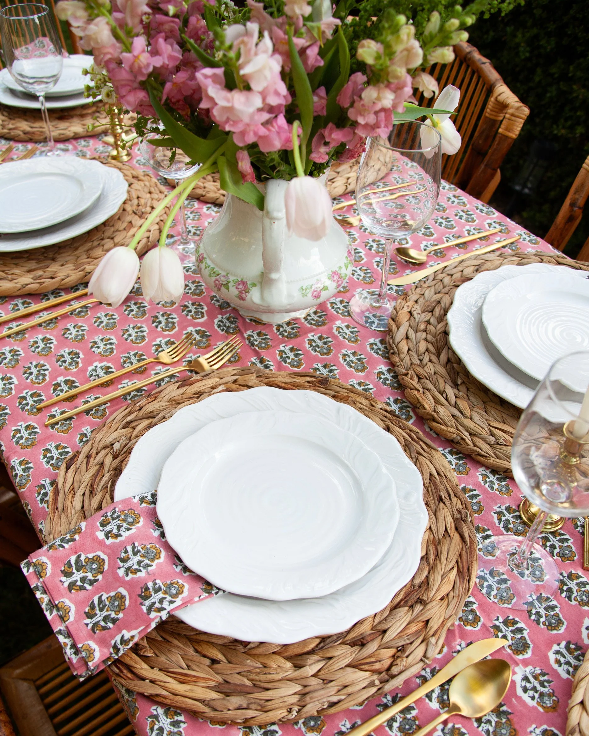 A table set for a meal with a floral centerpiece, white plates, gold flatware, clear wine glasses, woven placemats, and a pink floral tablecloth.