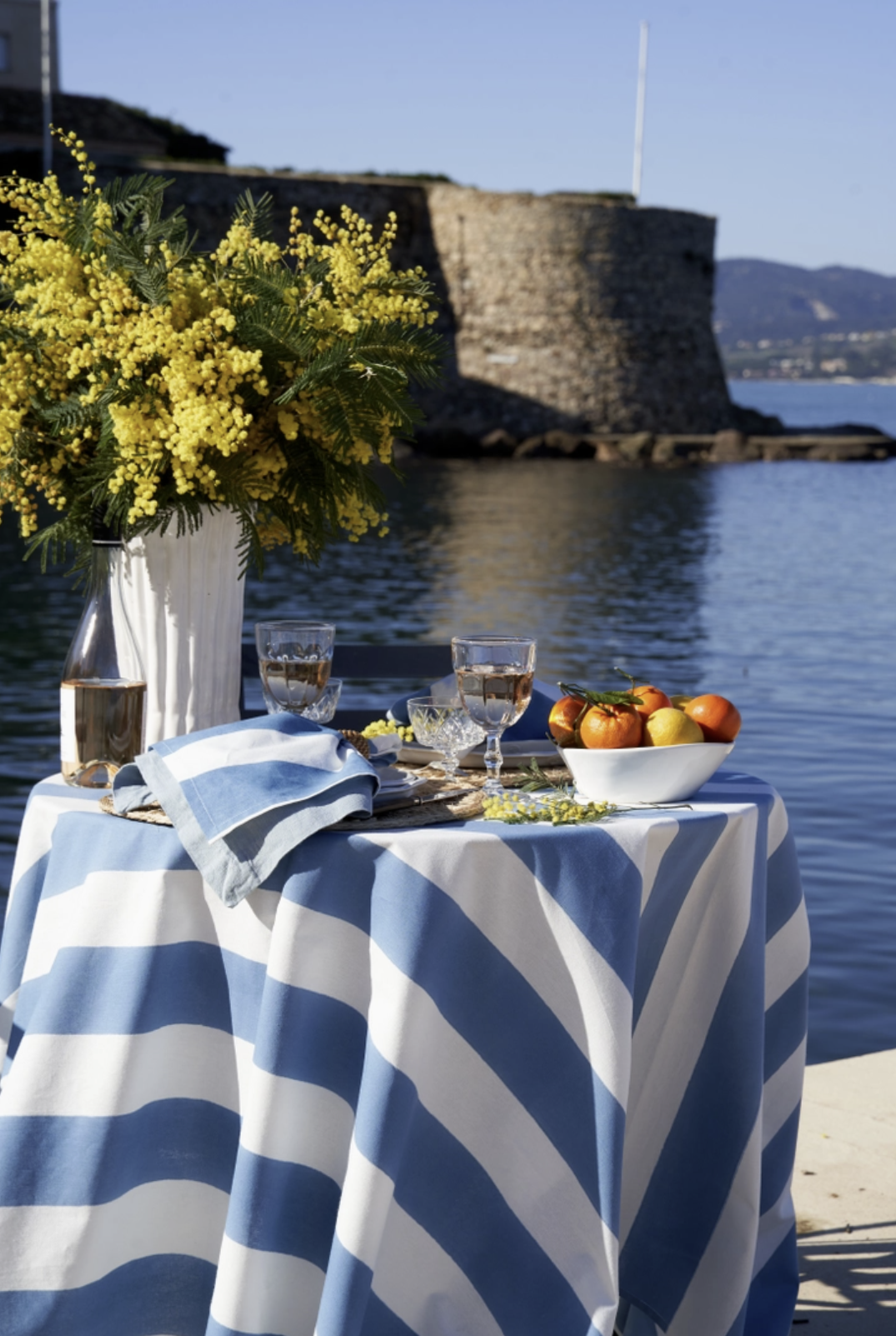 A table with a blue and white striped tablecloth set for a meal near the water, featuring a large yellow floral arrangement, glasses of water, a bowl of oranges and lemons, and a blue napkin, with a stone fort and water in the background on a sunny day.