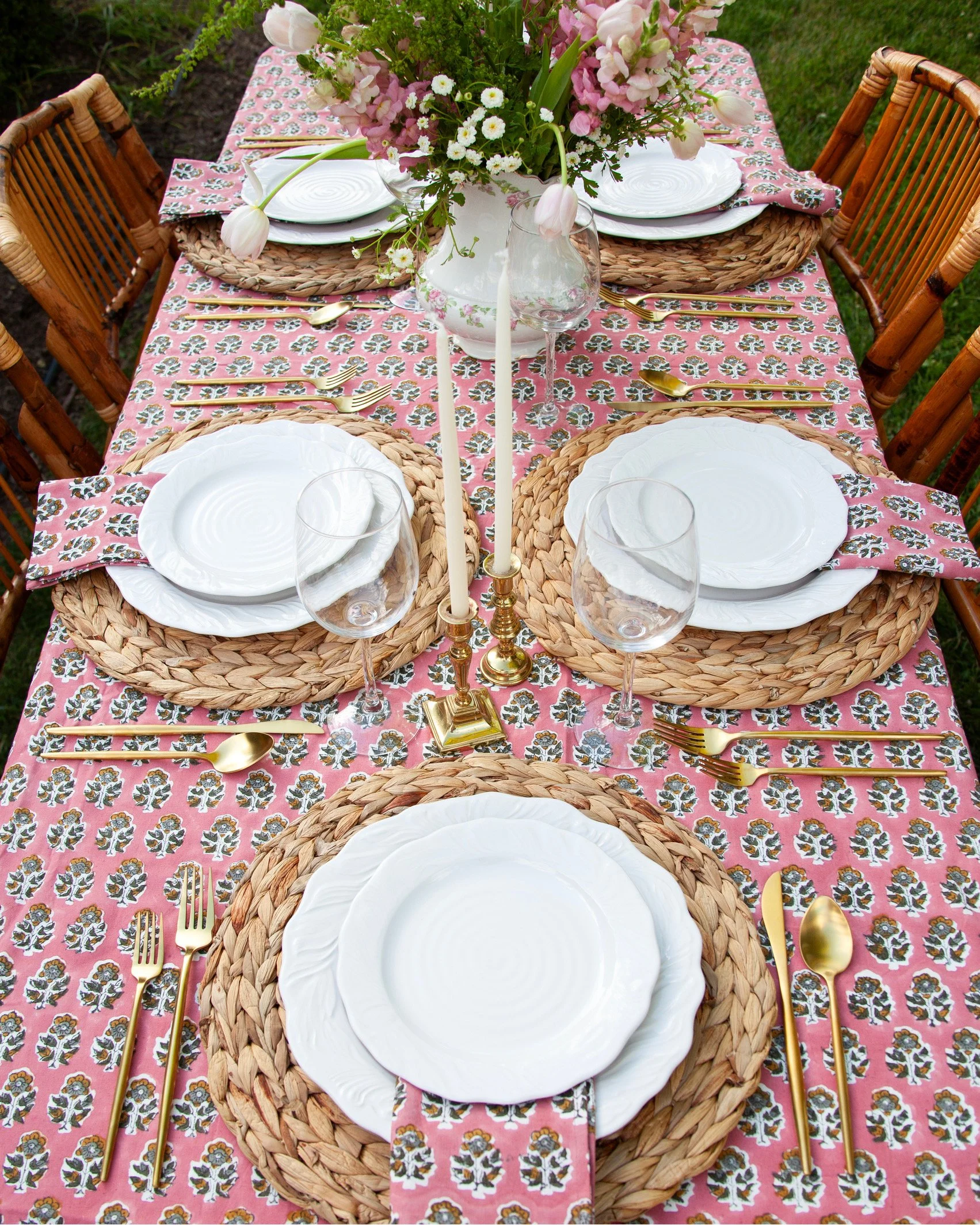 A close-up of a dining table set outdoors with a pink floral tablecloth, white plates on woven placemats, gold flatware, wine glasses, a floral centerpiece in a white vase, and tall taper candles in gold candle holders.