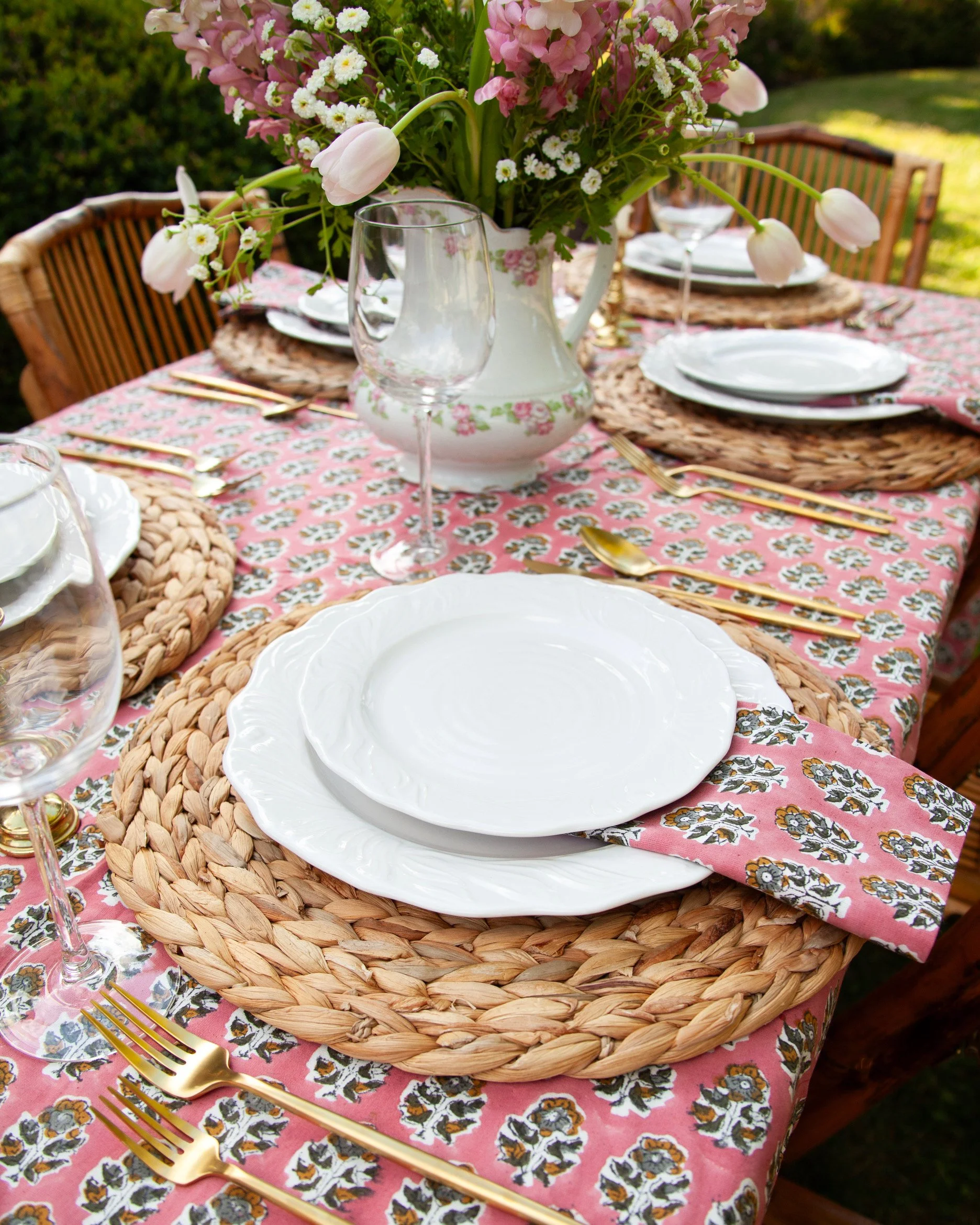 A table set for an outdoor meal with a pink floral tablecloth, white plates, gold flatware, and a large arrangement of pink and white flowers in a white pitcher at the center.