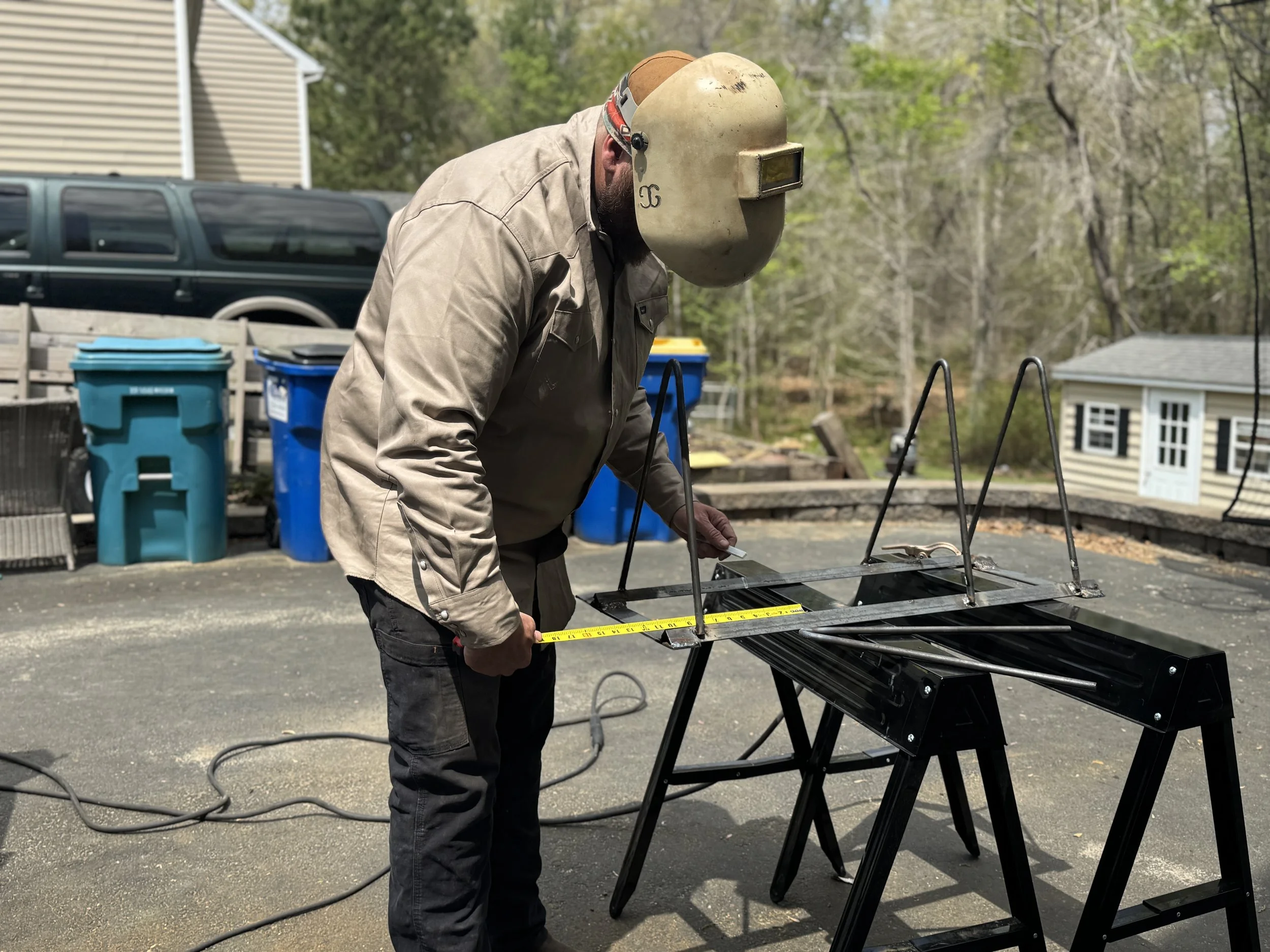 A man wearing a welding helmet and a beige work jacket is measured with a yellow tape measure while working outdoors at a welding or metalworking station.