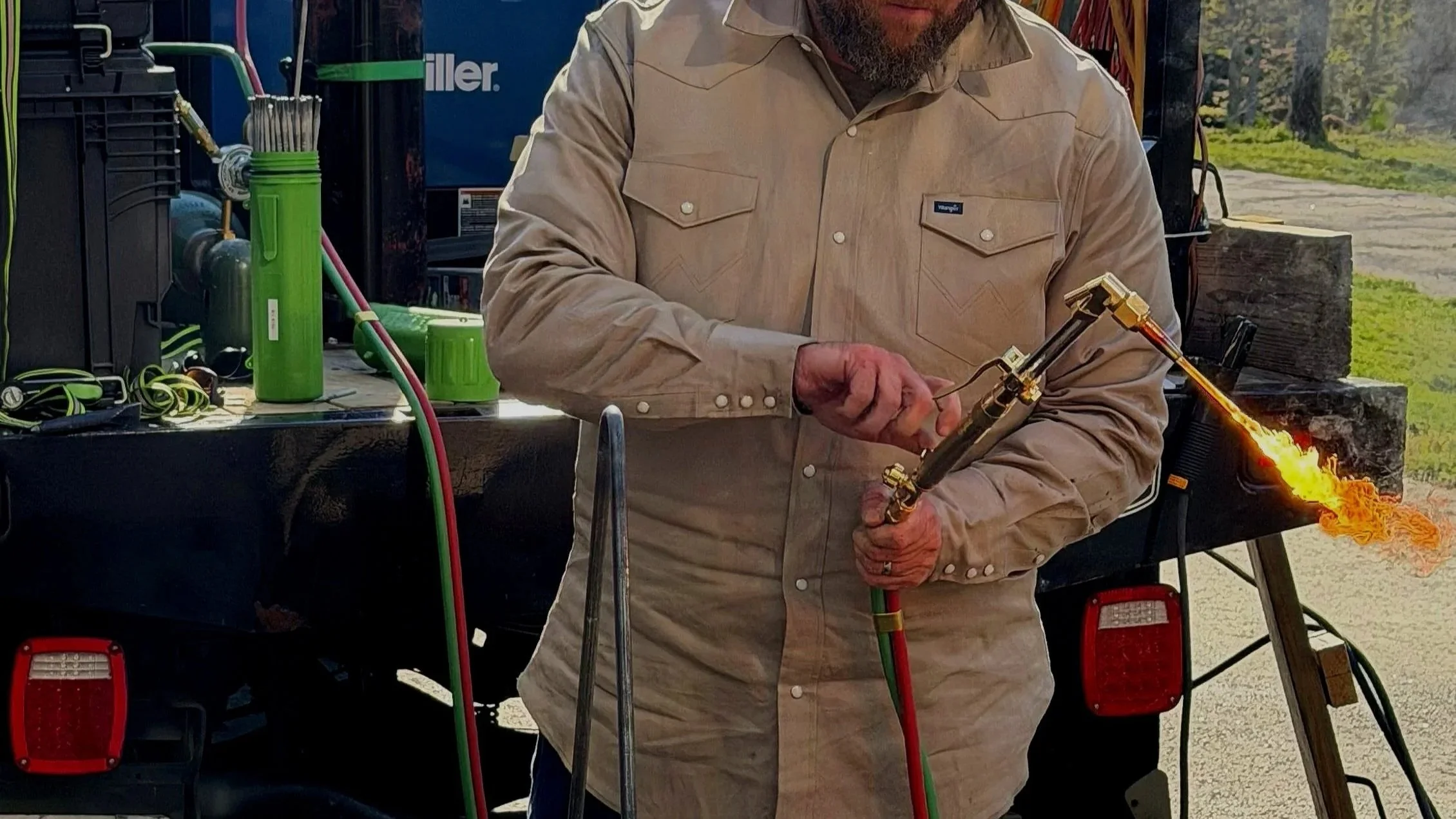 A man wearing a beige shirt holding a blowtorch with a flame, standing outdoors near a trailer with equipment and other items.