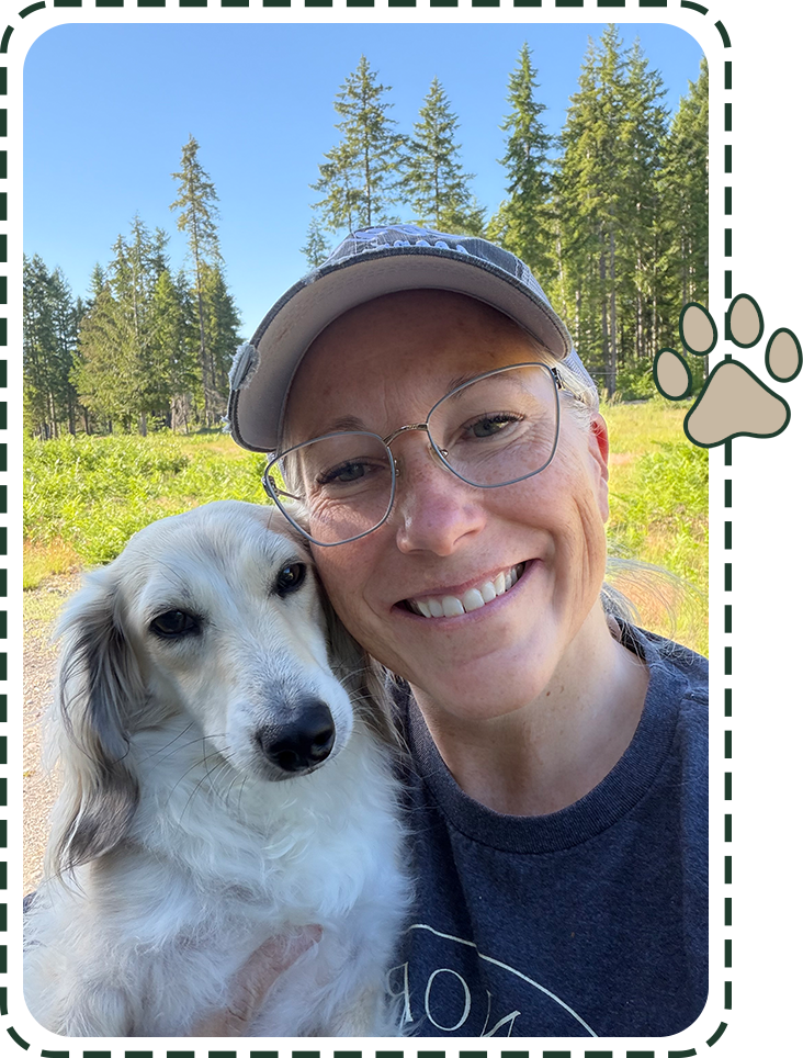 A woman smiling and holding a white and gray dog outdoors with green trees and blue sky in the background.