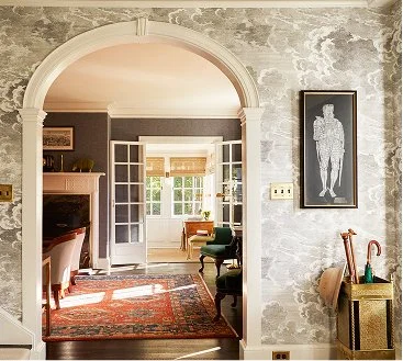 Living room view through a large arched doorway with sunlight, featuring a patterned rug, chairs, and a view of an interior space with windows and a small table.