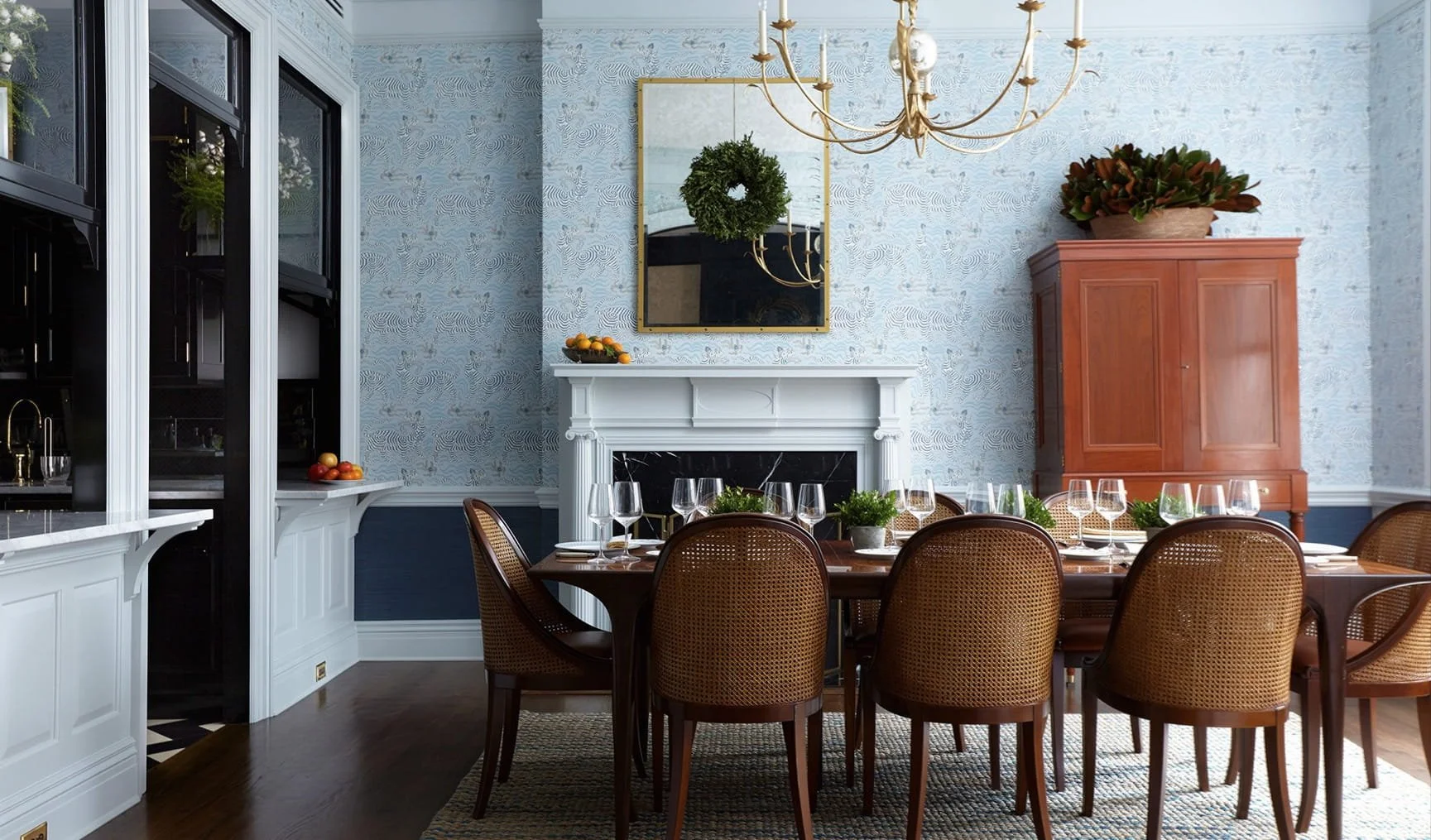 A decorated dining room with a large wooden table set with wine glasses and small potted plants, surrounded by brown woven chairs. There is a white fireplace with a mirror and green wreath above it, a wooden cabinet with a plant on top, and a chandel