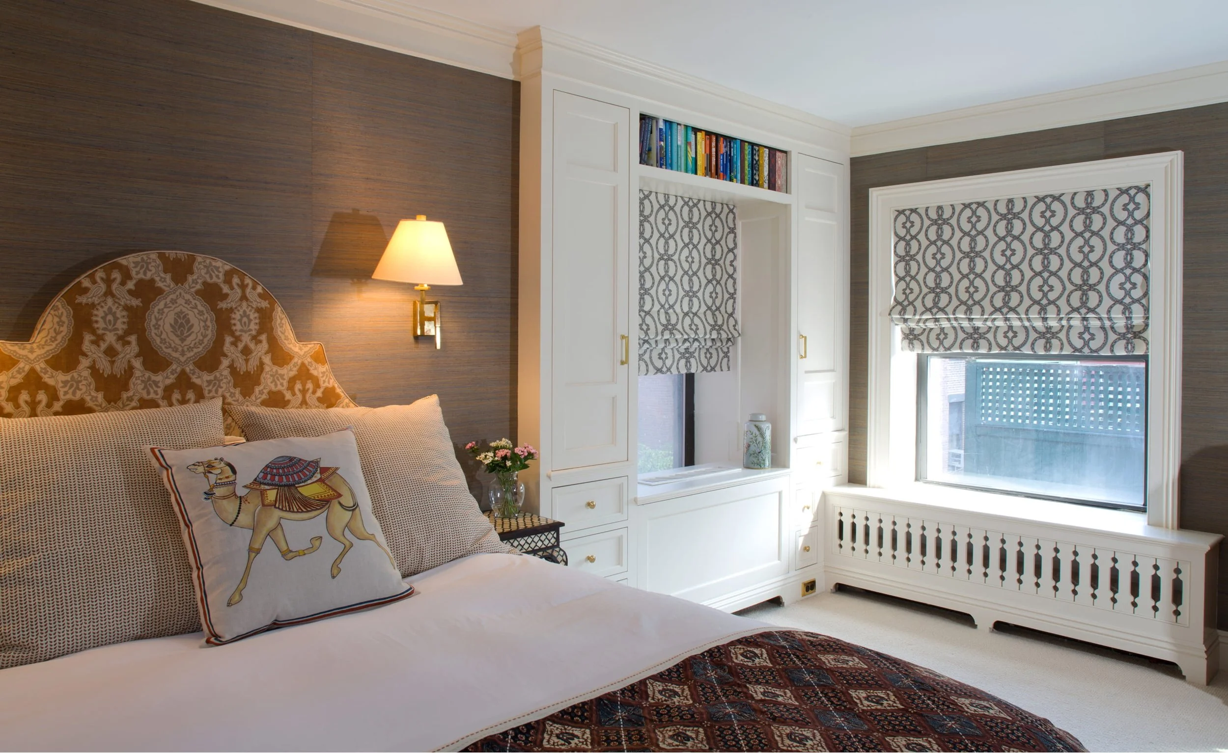 A bedroom with a bed featuring decorative pillows, a wall-mounted lamp, a small vase with flowers on a side table, built-in white shelves with books, and windows with patterned Roman shades. The room has a mix of dark and light decor elements.