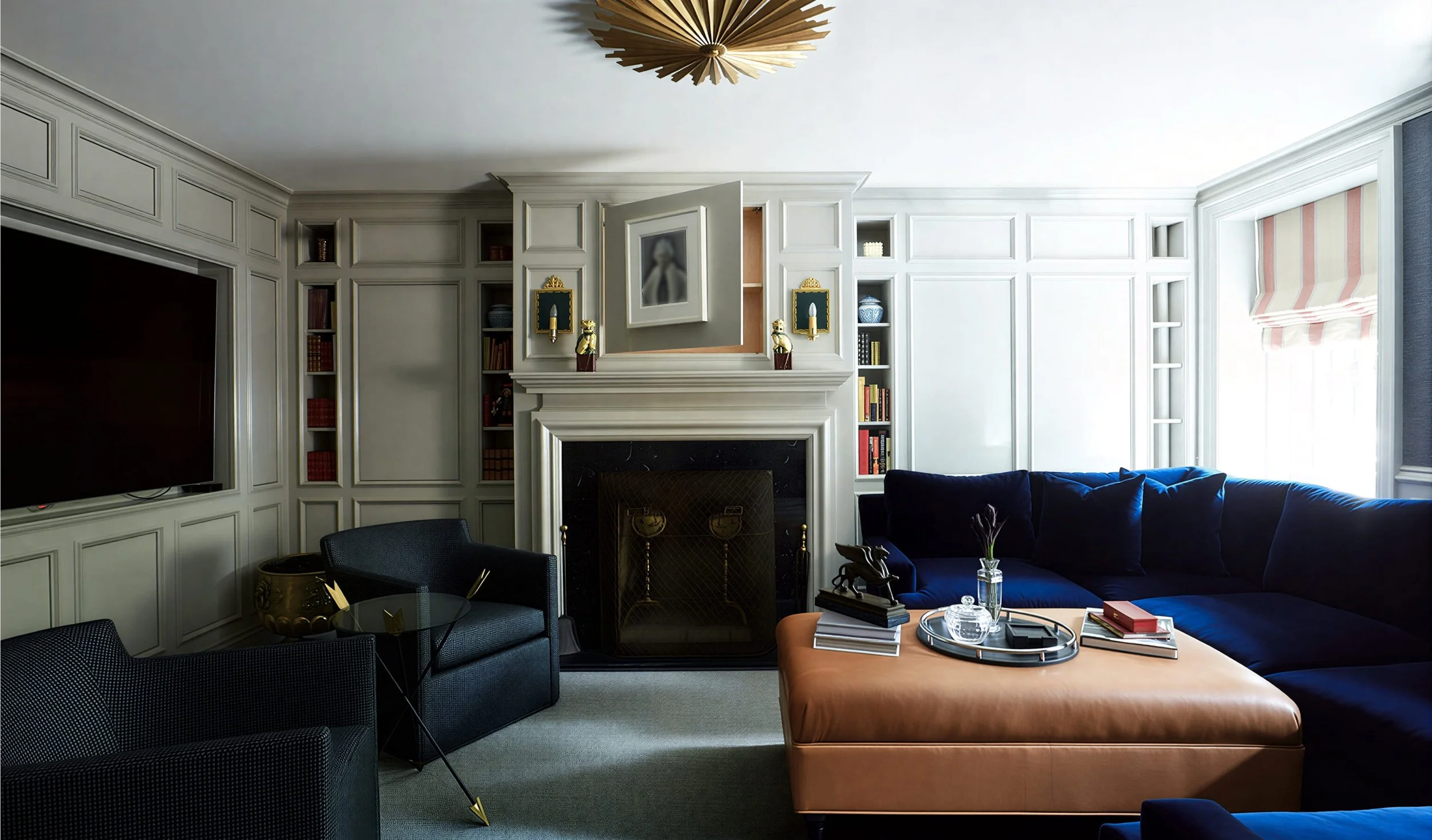 Living room with white paneled walls, built-in shelves, and a decorated fireplace mantel. There is a navy blue sectional sofa, a tan ottoman, and a small round table with books and decorative items. A black armchair and a glass-top side table are als