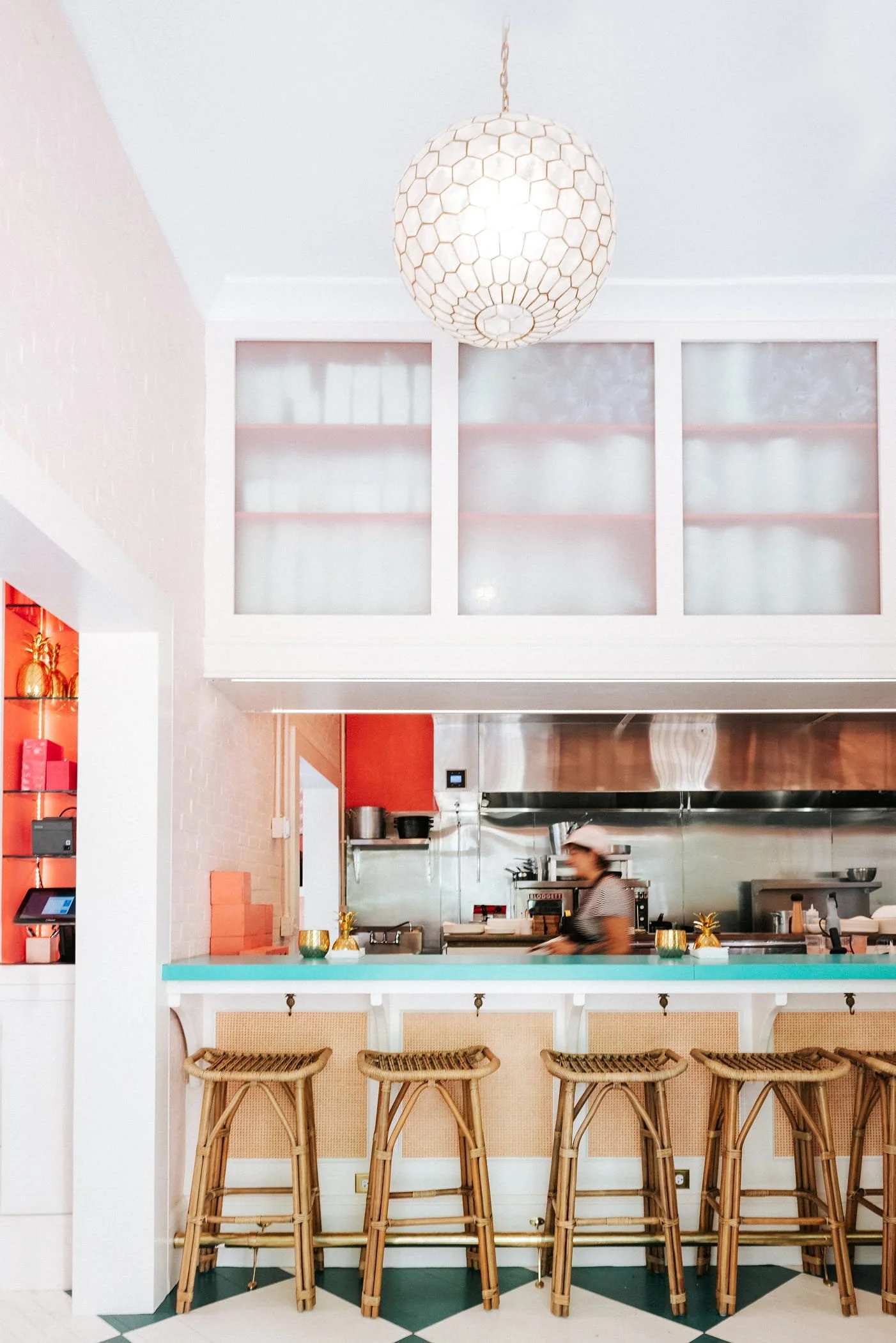 A restaurant or cafe interior with a light pink and white color scheme, a row of high wooden bar stools at a teal counter, and a kitchen area visible in the background with a person working.