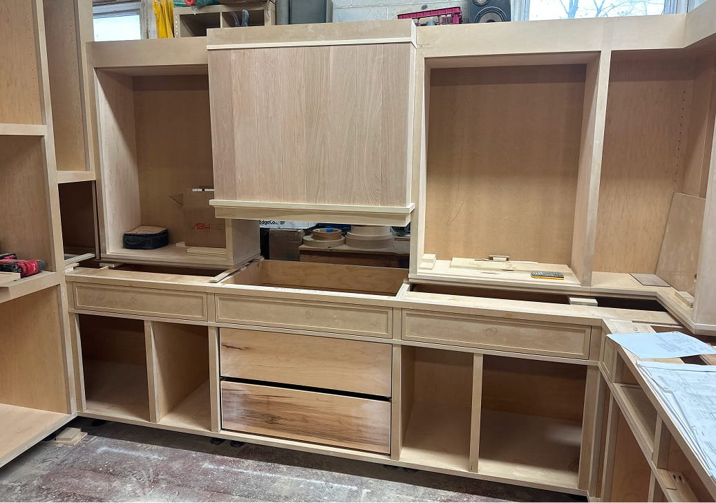 Unfinished wooden kitchen cabinetry with drawers and open shelves in a workshop.