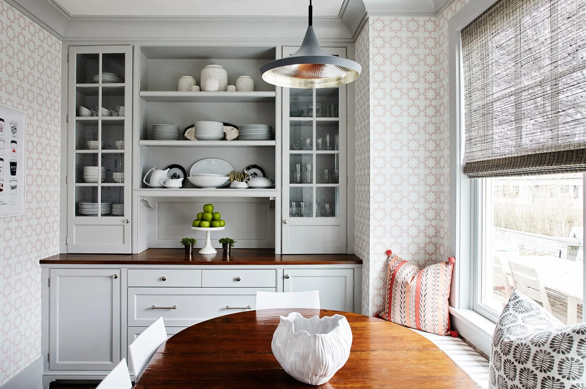 Kitchen corner with white cabinetry and open shelving holding white dishes and glassware, a wooden table with a decorative white vase, and a window with woven blinds and patterned cushions.