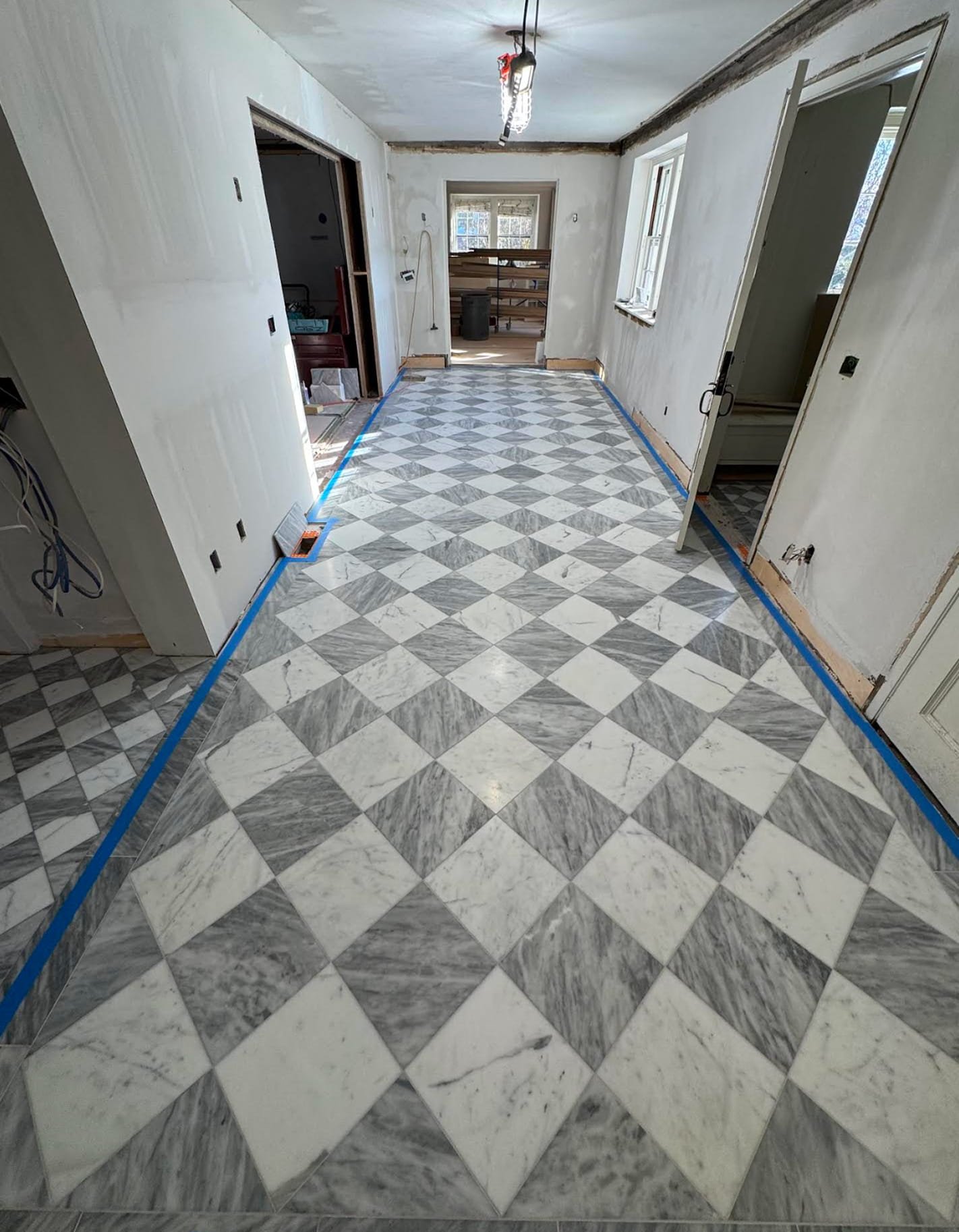 Interior room under renovation with marble patterned tile floor, partially finished white walls, and ceiling. Blue painter's tape outlines the floor edges, and a light fixture is attached to the ceiling. Windows on the right wall and an open doorway at the back show additional space.