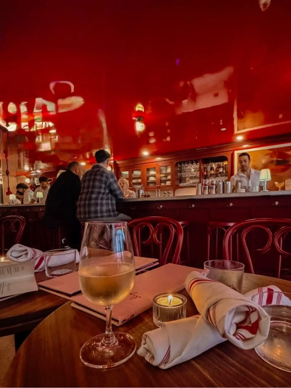 A cozy restaurant with tables set for dining, including a glass of white wine and rolled napkins on a wooden table, and a bar area in the background with patrons and a bartender.