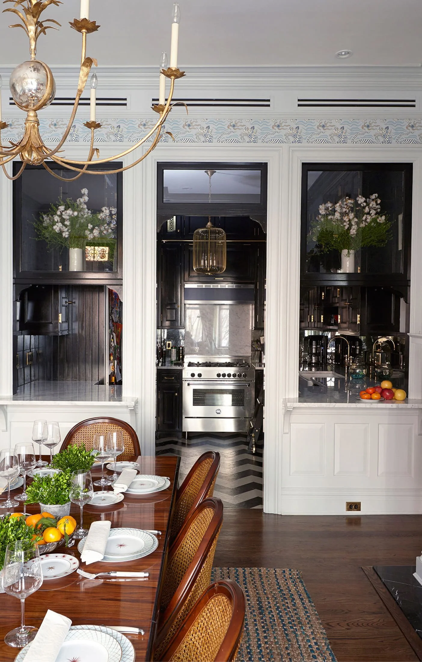 Elegant dining room with a wooden table set for dinner, featuring white plates, wine glasses, and napkins. A centerpiece with greenery and oranges is on the table. The background shows a kitchen with dark cabinets, a stainless steel stove, and decorative vases with white flowers on top of a black built-in shelf. A chandelier with gold accents hangs from the ceiling.