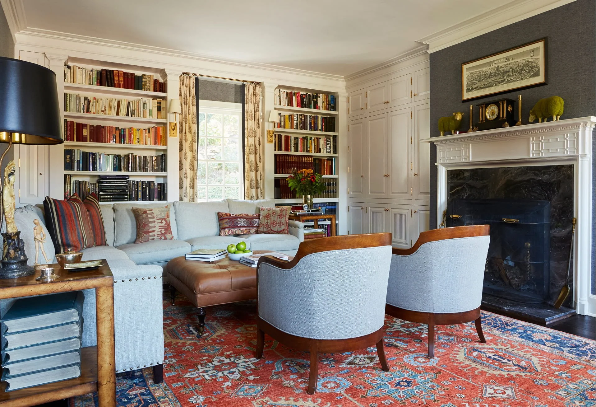 A cozy living room with a white sofa, colorful pillows, and a glass coffee table with green apples, surrounded by chairs and a vintage rug. There are built-in bookshelves filled with books, a window with floral curtains, a fireplace with decorative items, and a framed picture above it.
