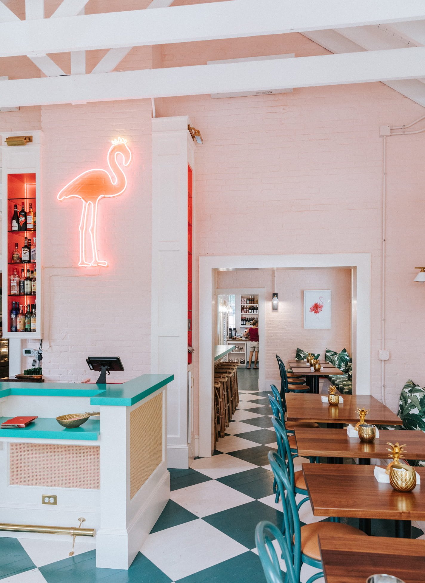 Interior of a restaurant with pink brick walls, a neon flamingo sign, and a dining area with wooden tables, tropical-patterned chairs, and gold pineapple decorations.