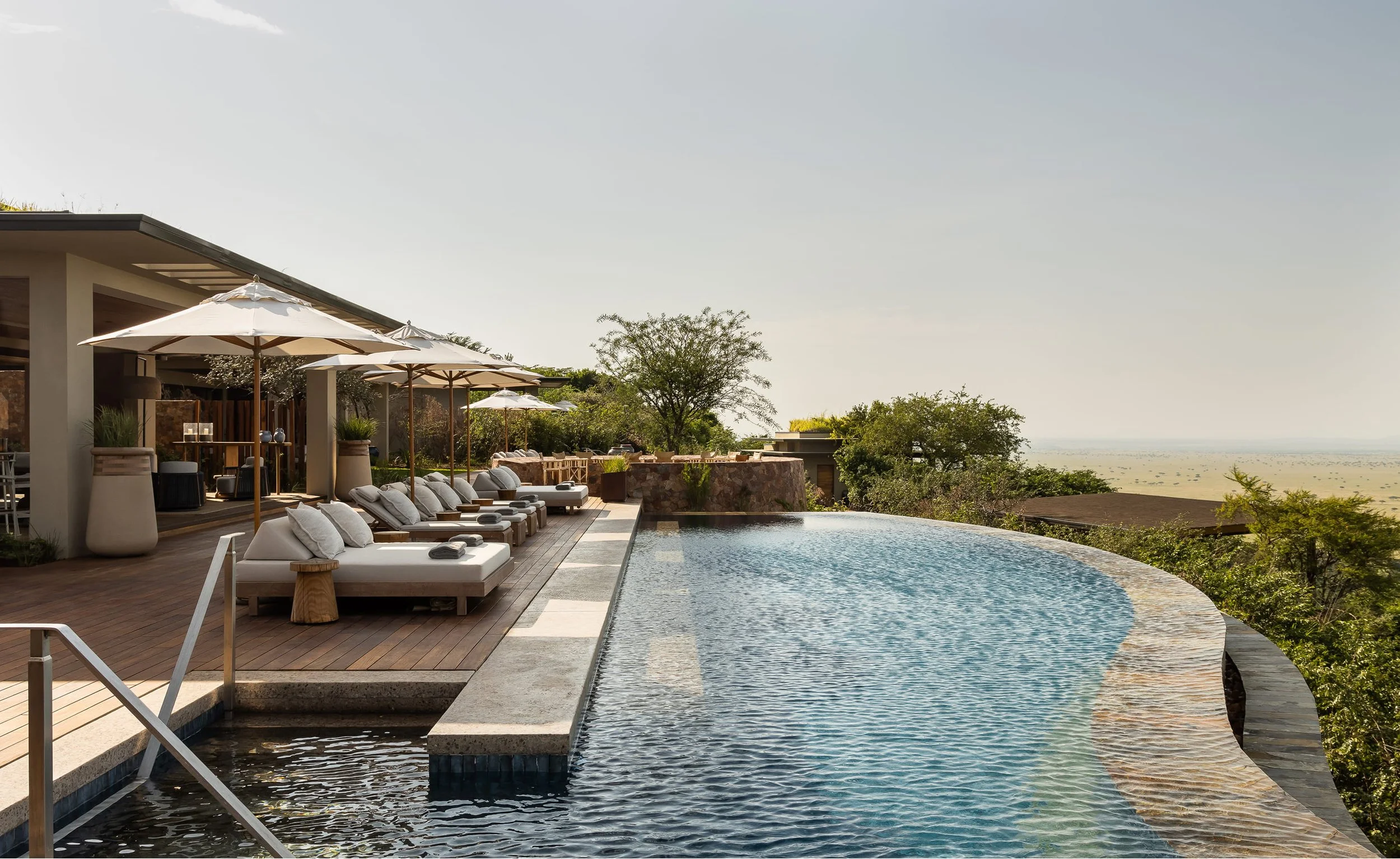 Luxury outdoor infinity pool with lounge chairs and large white umbrellas at a modern house on a hill, overlooking a distant landscape and sky.