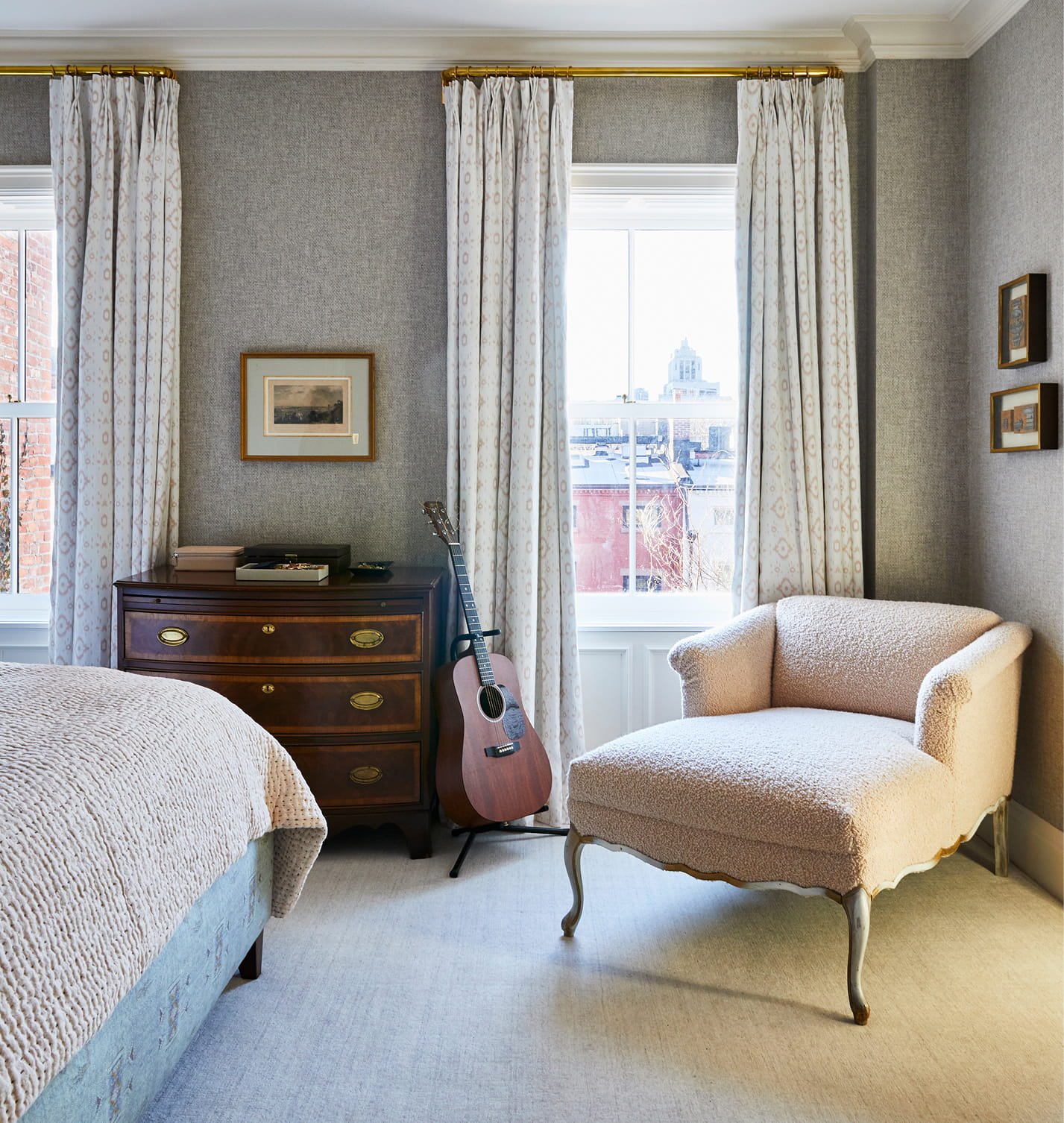 A cozy bedroom corner with a window, white curtains, a vintage wooden dresser, an acoustic guitar, a plush cream-colored armchair, and a bed with a textured beige blanket.