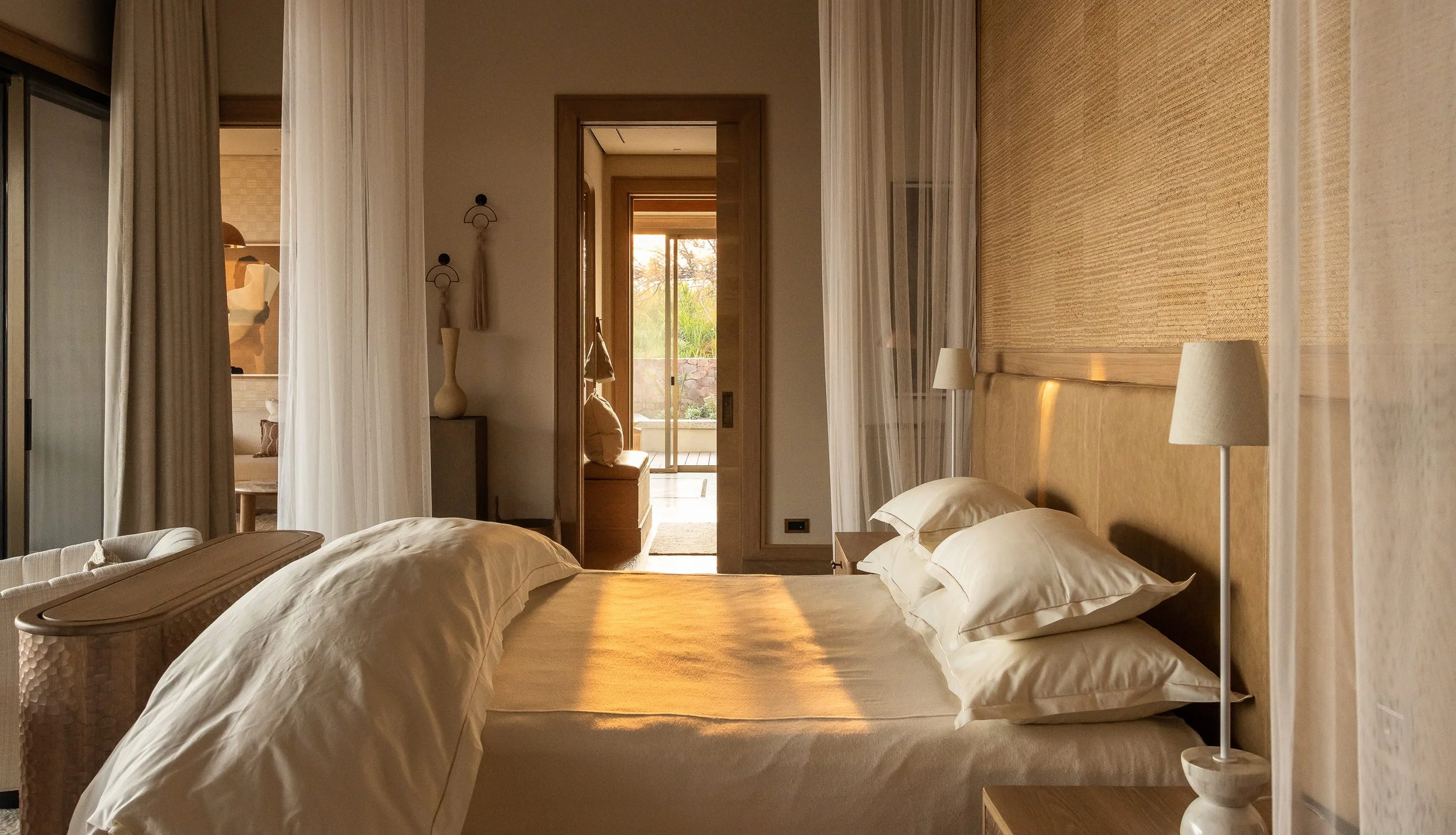 A cozy bedroom with a large bed, white bedding, and two pillows. There are nightstands with lamps on each side of the bed. A sliding glass door at the foot of the bed leads to a balcony or patio with greenery outside. The room has warm lighting and neutral, earthy tones.
