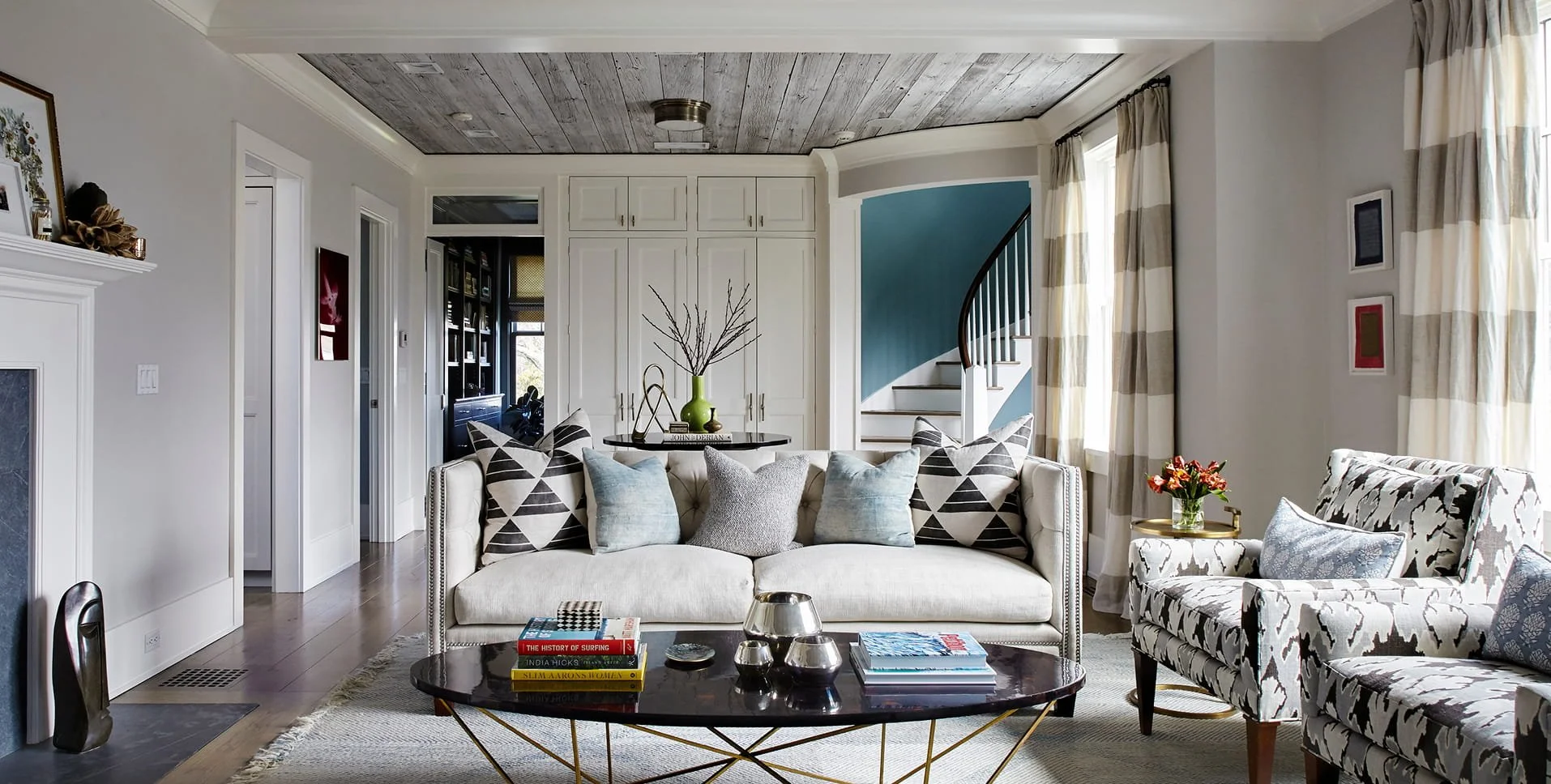 Bright living room with white walls, patterned furniture, a black coffee table with books and decor, and a staircase with a blue wall in the background.