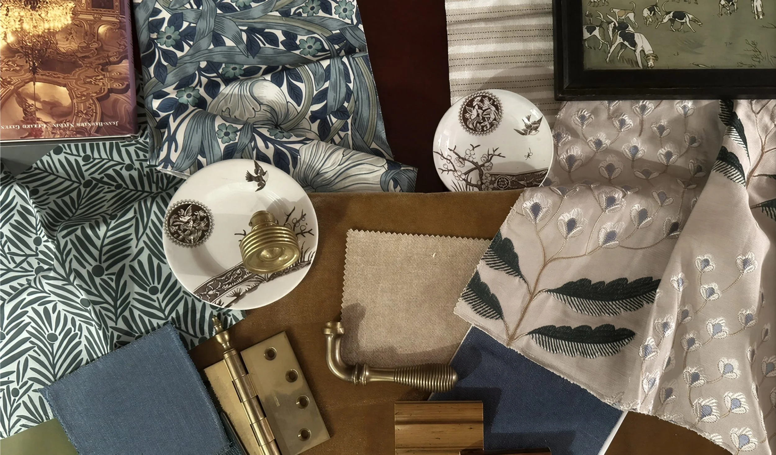 Display of various fabric samples with patterns including floral, leaf, and abstract designs, alongside a small white decorative plate with bird and branch motifs, a brass curtain rod, and a brass hinge.