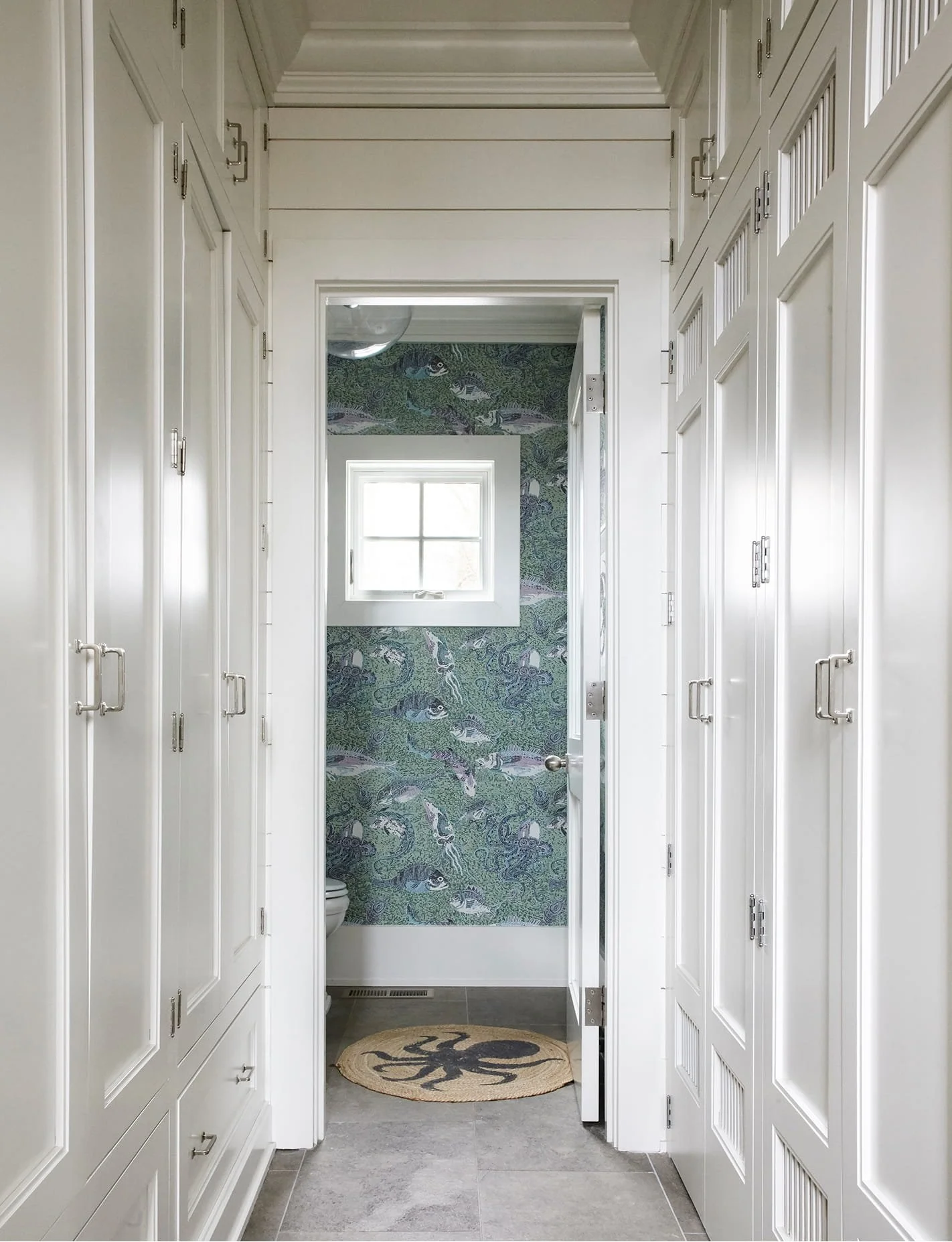 White hallway with built-in cabinets and closet doors on both sides, leading to a small bathroom with fish and aquatic plant wallpaper, a window, and a round rug with a black octopus design.