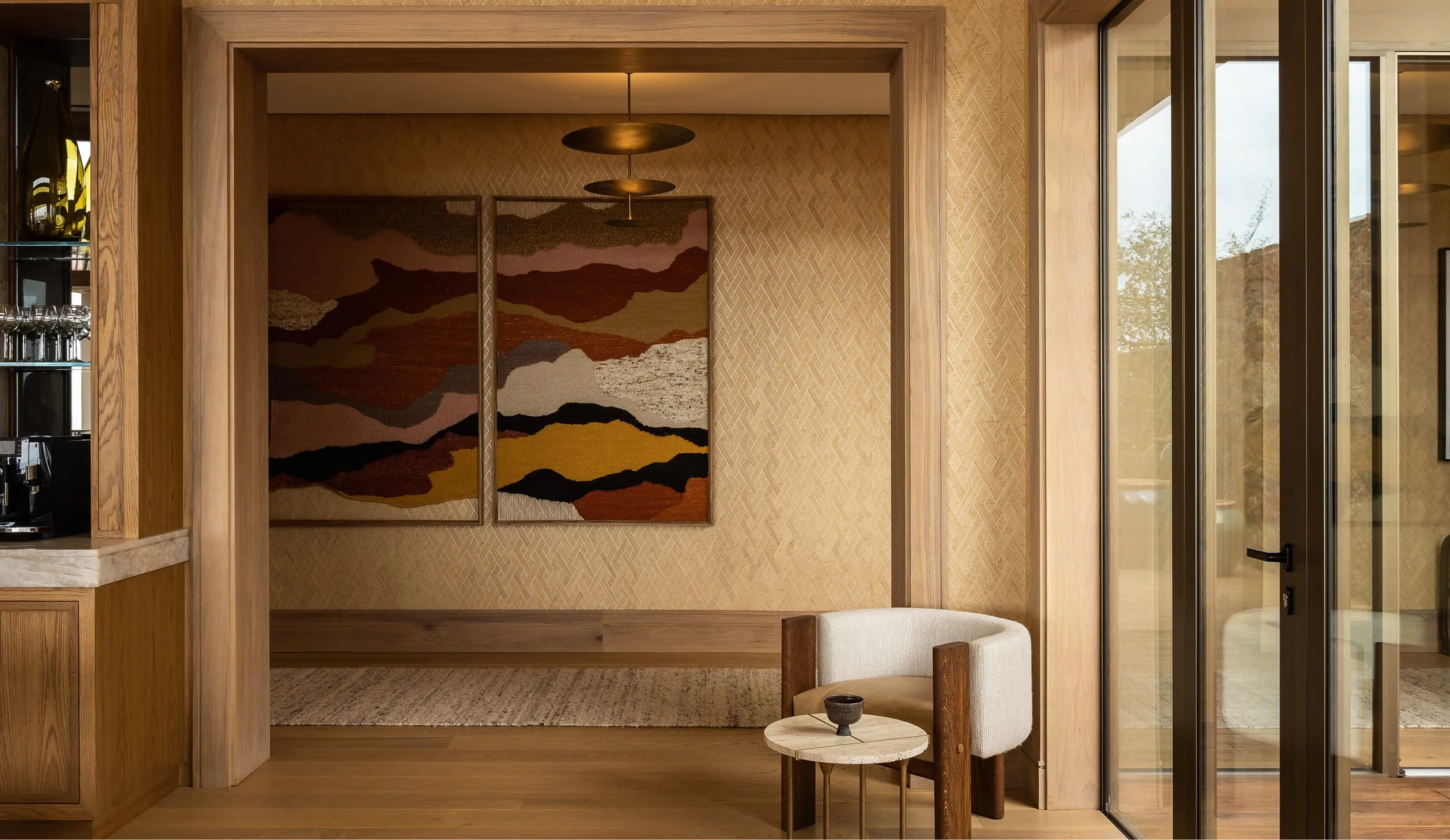 Interior view of a room with wooden framing, a large colorful abstract art piece on the wall, and a glass door to the right. There is a chair and small round side table in the foreground.