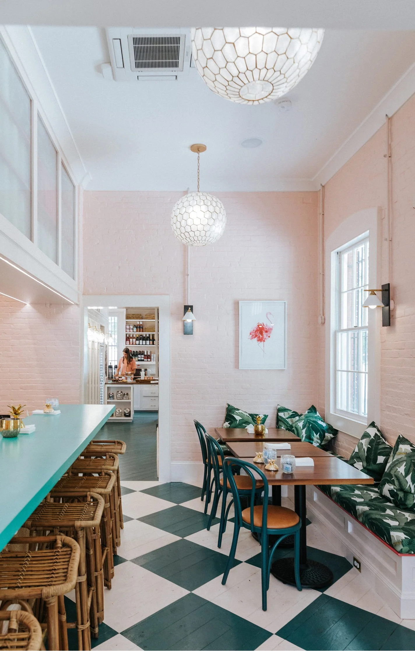 Interior of a bright cafe with white painted brick walls, green checkered floor, wooden tables with green and leaf patterned cushions, and wicker barstools. Decor includes a framed flamingo print, hanging light fixtures, and a bar area in the background with shelves of bottles.