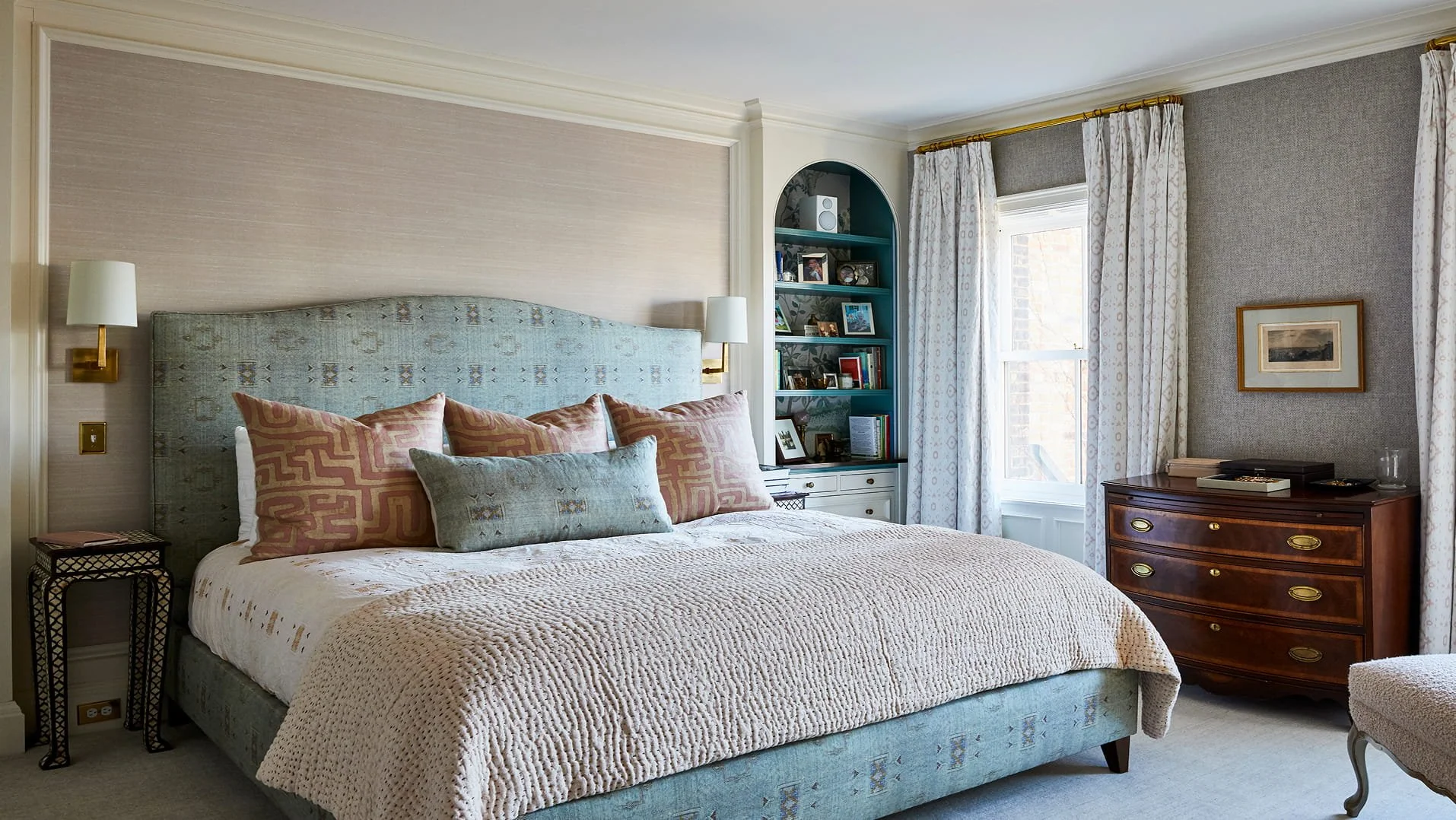 A cozy bedroom with a large bed, patterned pillows, and a light-colored textured bedspread. The room features a blue upholstered headboard, wall-mounted lights on either side of the bed, a nightstand, a built-in bookshelf with framed photos and books, a window with patterned curtains, and a wooden dresser with framed artwork above it.
