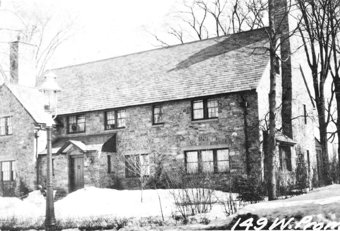 Black and white photo of a stone house with a steep roof, several windows, a wooden door, bare trees, and snow on the ground.