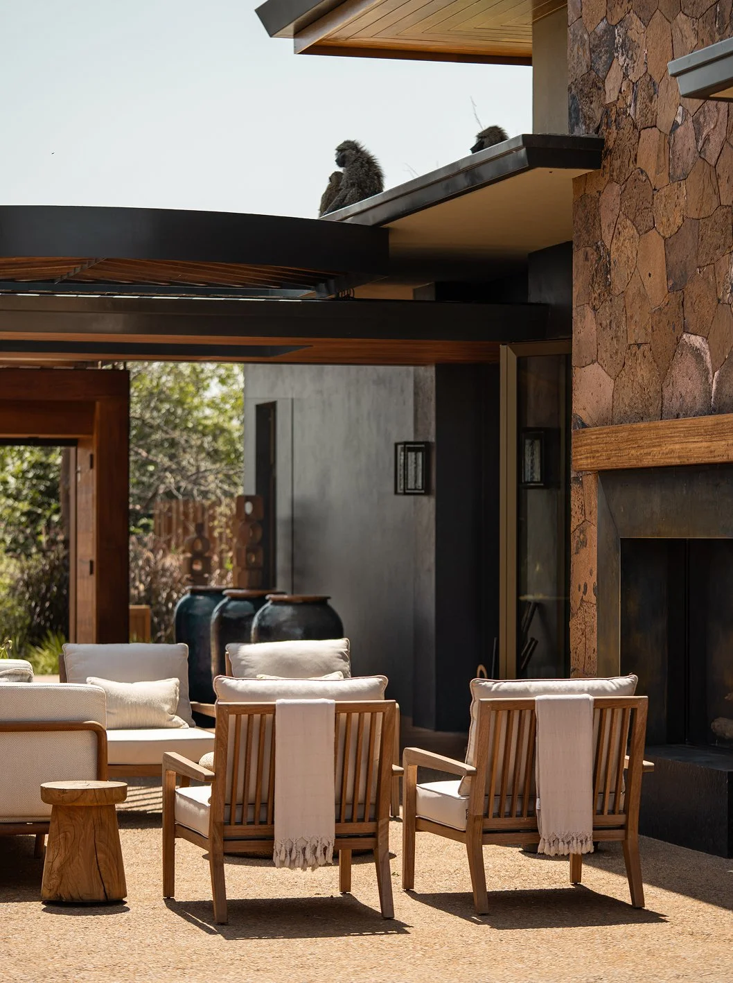 Outdoor patio with wooden chairs, white cushions and throws, a stone fireplace, and a modern house with large windows and black and wooden accents.