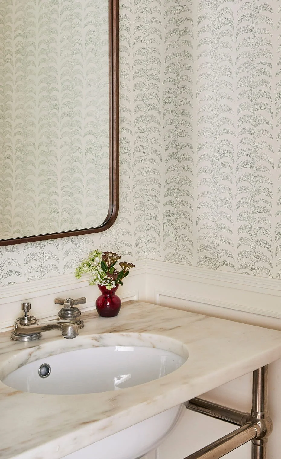 Bathroom sink with marble countertop, a small red vase with flowers, a mirror above the sink, and patterned wallpaper in the background.