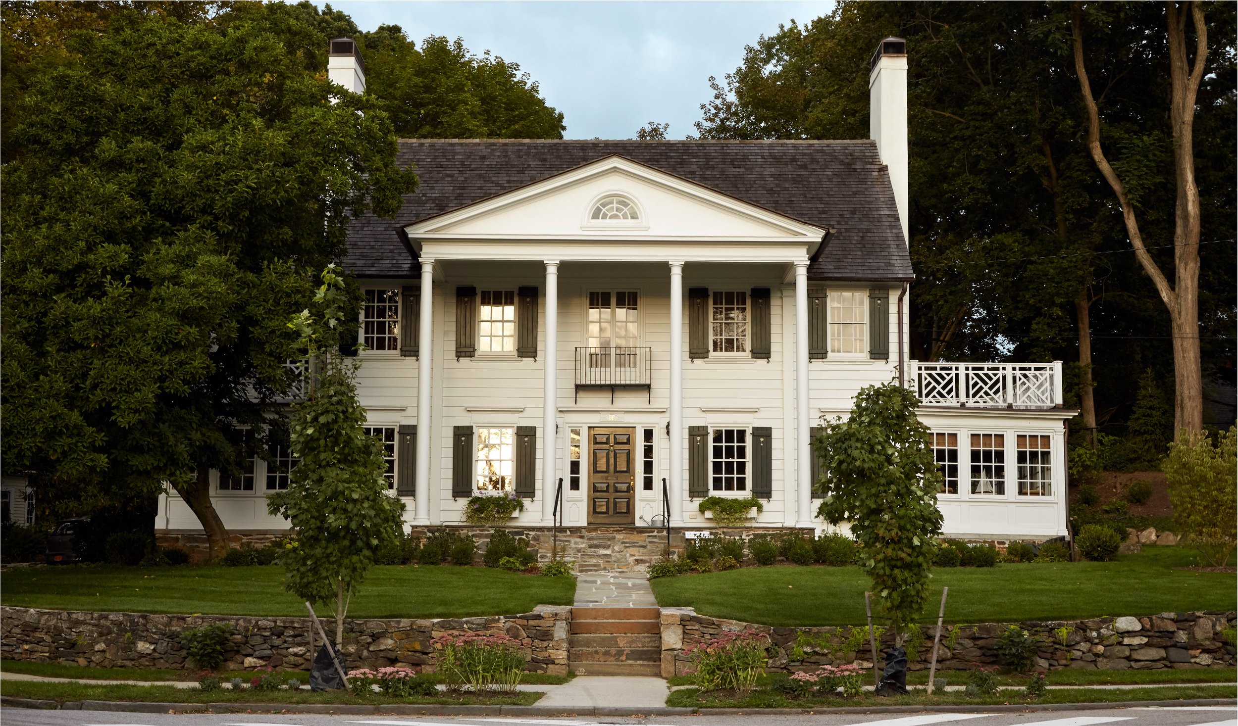 A large, white, two-story house with black shutters, a front porch with columns, and a balcony on the right side. The house has a stone foundation, and there are new trees and plants in the front yard, with a stone pathway leading to the front door.