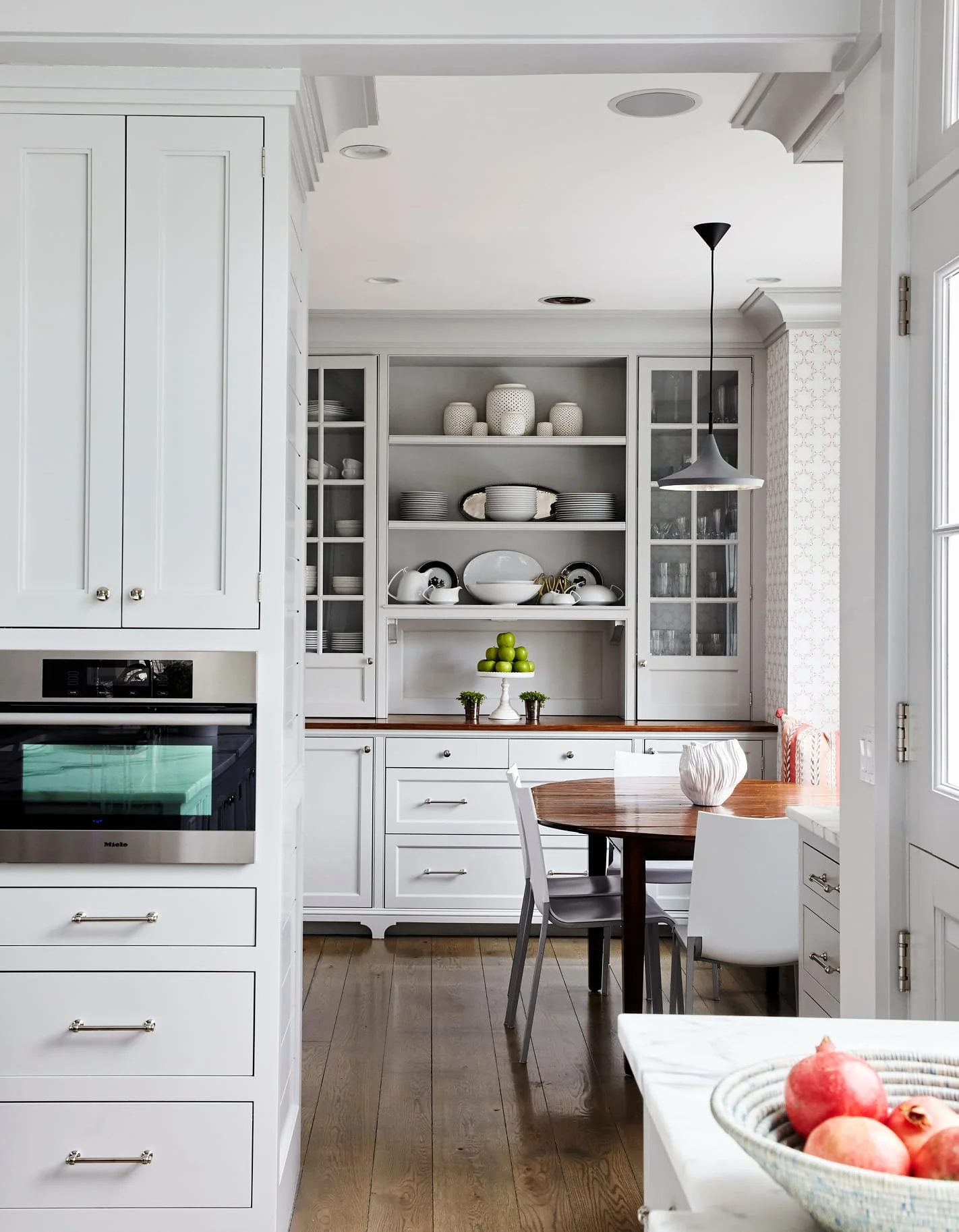 White kitchen with open shelves displaying dishes, a small table with a white chair, a fruit bowl with pomegranates, and a dining table with a white decorative vase.