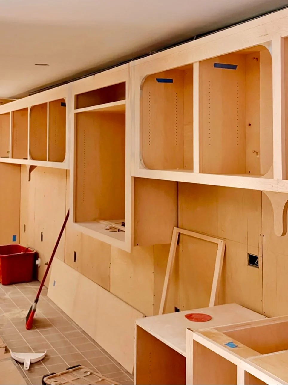 Wooden cabinetry being installed in a room, with tools like a broom and dustpan on the tiled floor.