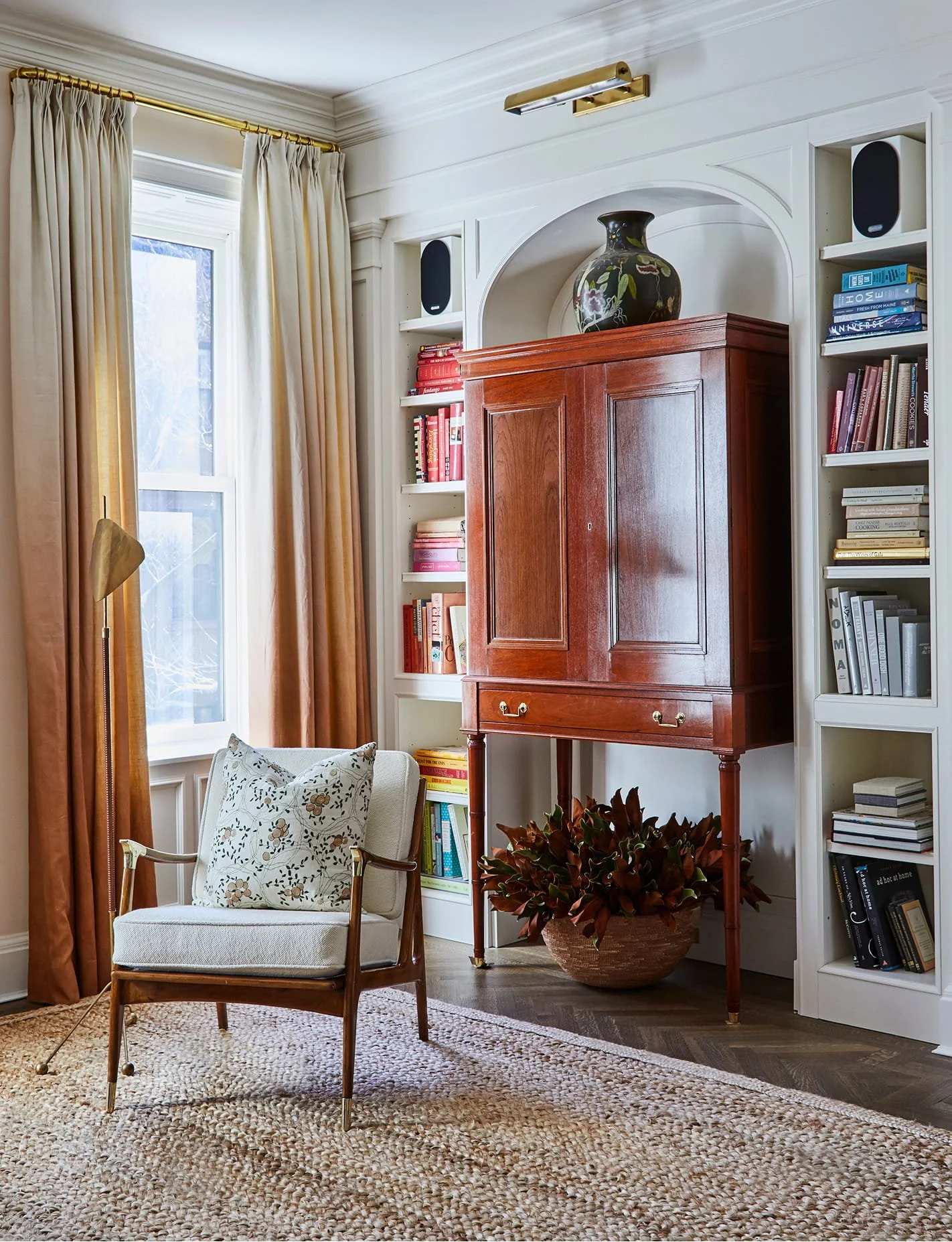 Living room corner with large window, cream curtains, white bookshelf filled with books, a wooden cabinet, potted plant, armchair with floral pillow, and a woven rug.