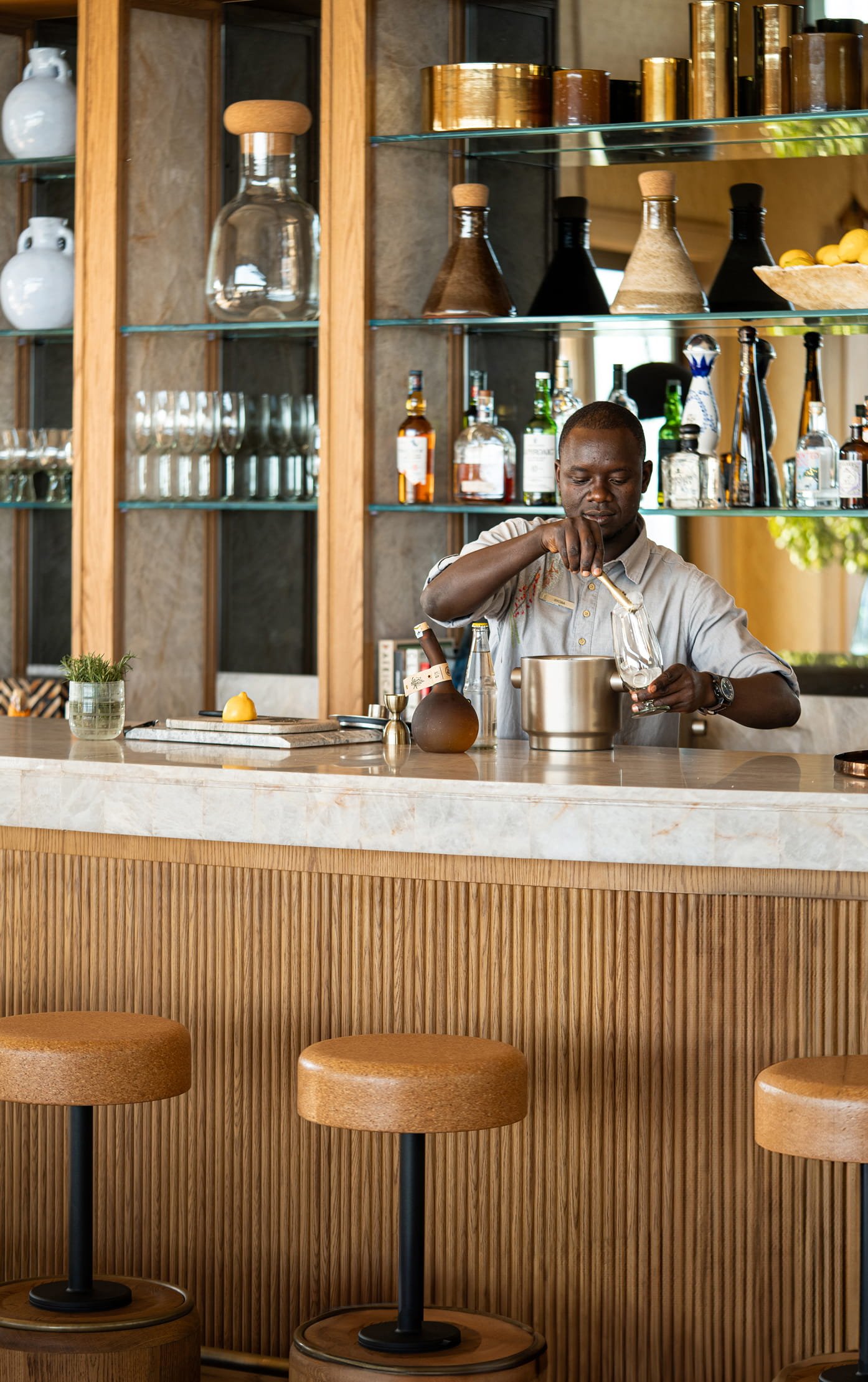 A bartender pouring a drink in a glass at a wooden bar counter with bar stools, bottles, and glassware behind him.
