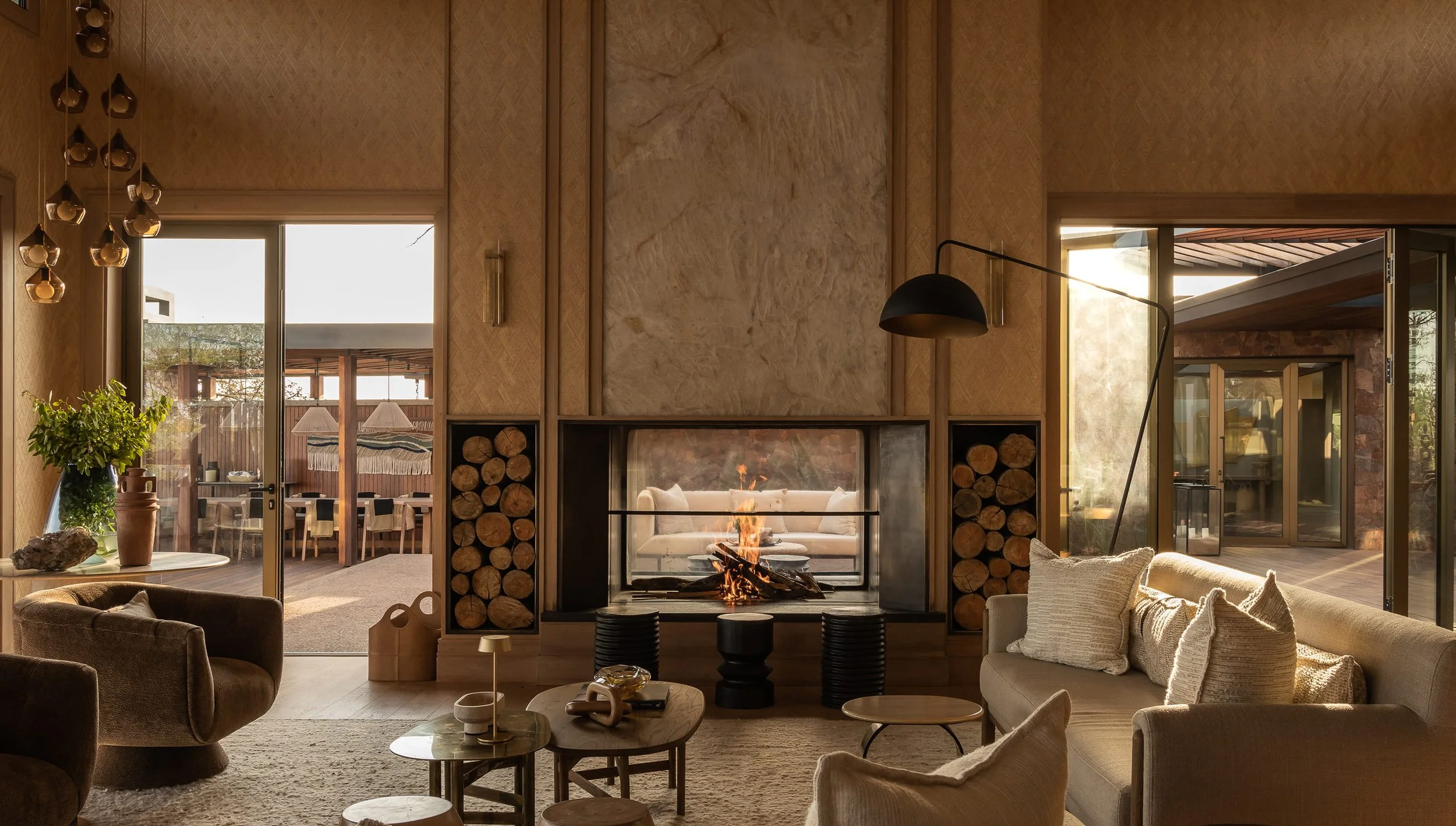 Living room with a fireplace, wood logs on both sides, beige sofas, a black arc floor lamp, and large sliding glass doors leading outside.