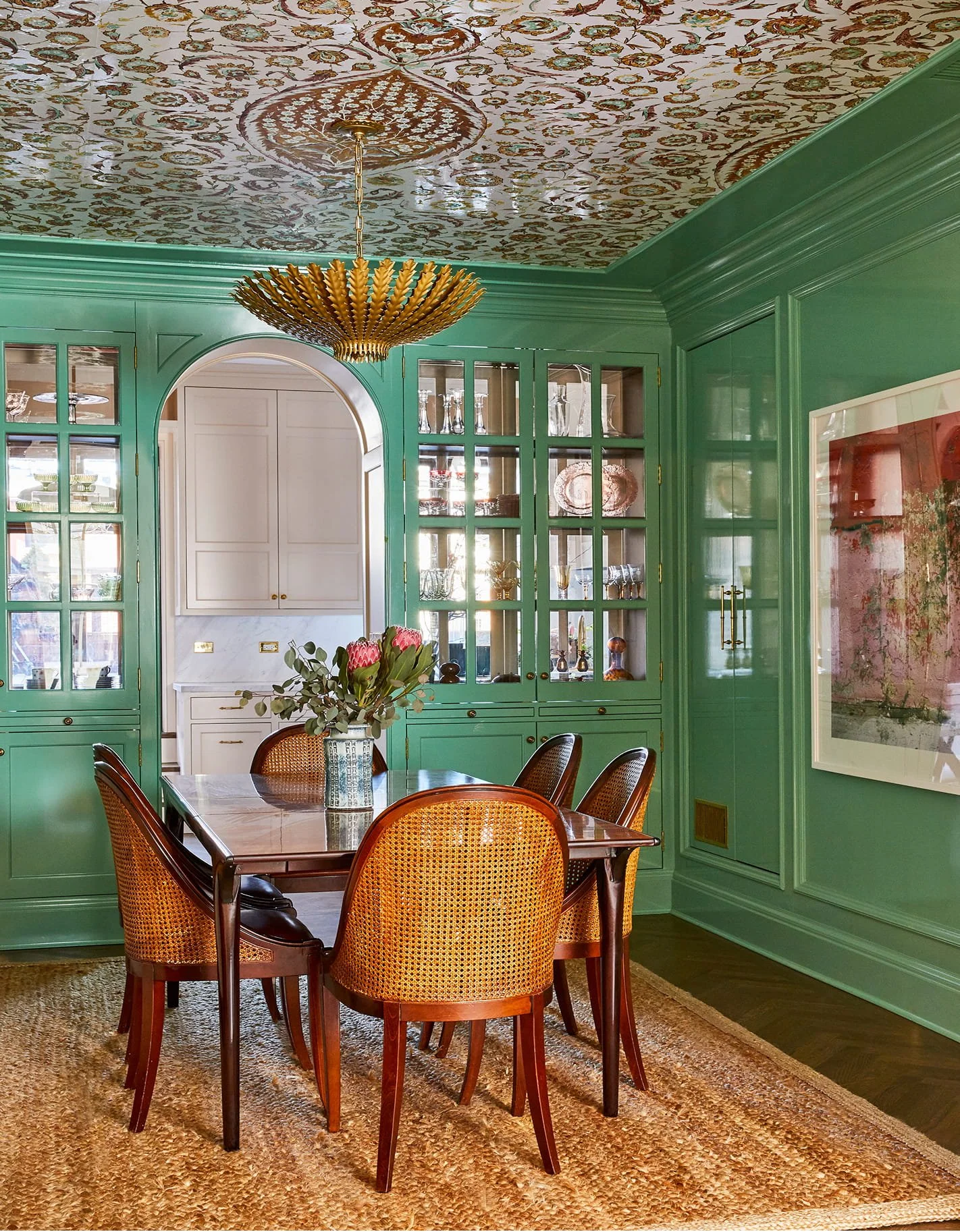 A dining room with green paneled walls, a wooden table, and six woven-back chairs. A gold chandelier hangs from a decorated ceiling, and a bouquet of flowers on the table.