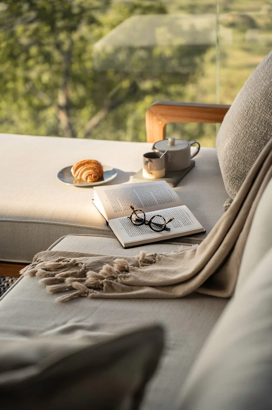 A cozy outdoor lounge setup with an open book, a pair of glasses, a blanket, a croissant on a plate, and a tea set on a table, with a view of green trees in the background.