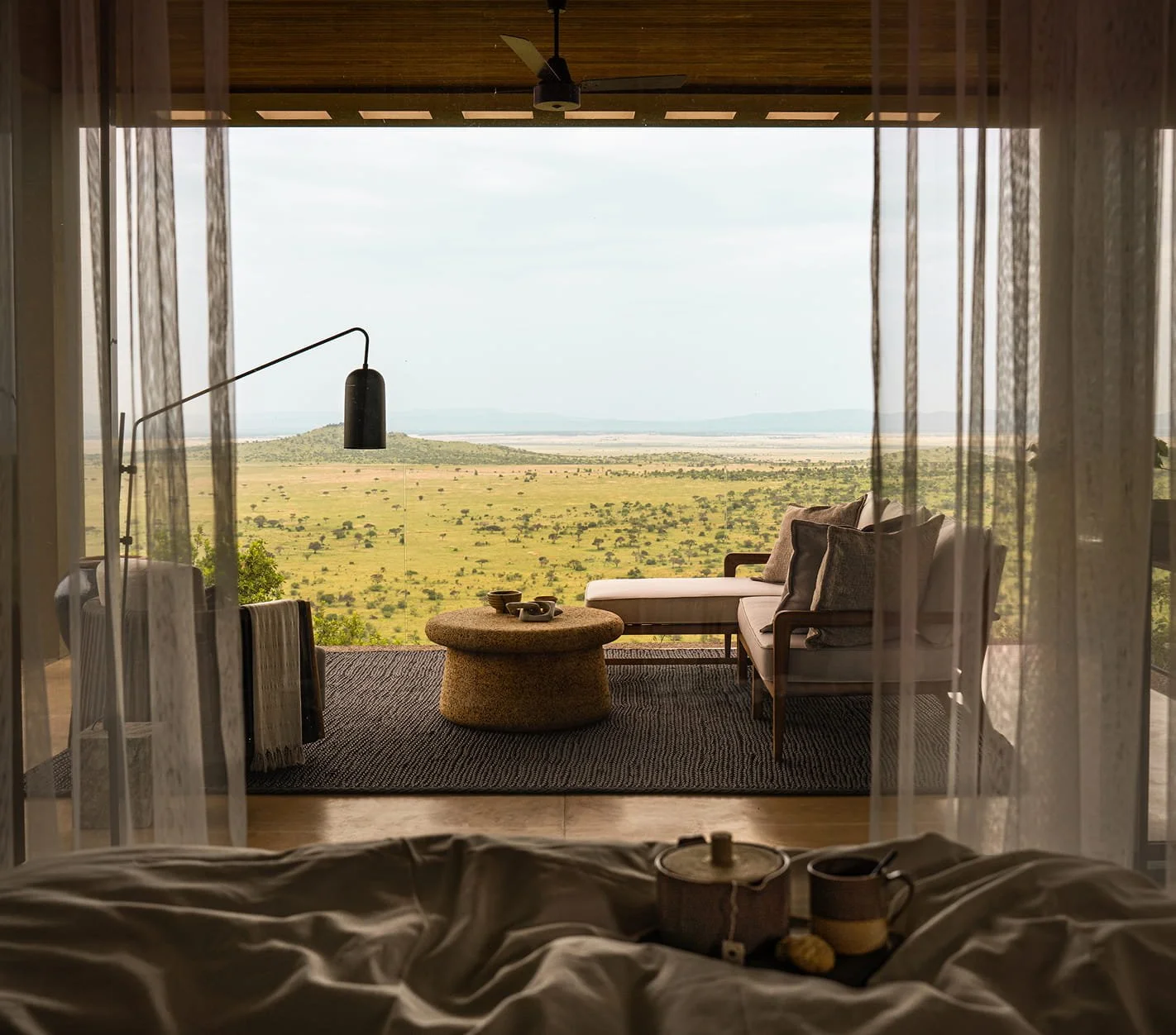 View from an indoor bedroom looking out through sheer curtains onto a balcony with outdoor furniture and a vast savannah landscape with a small hill in the distance.