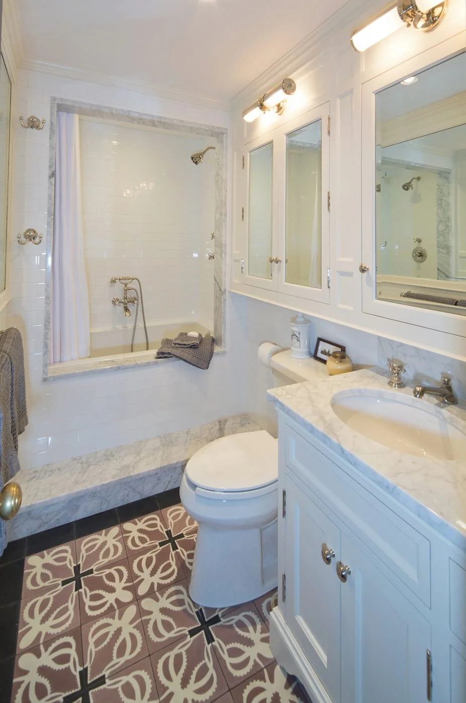 A small, elegant bathroom with a bathtub and shower, white subway tiles, a mirrored medicine cabinet, white cabinetry with a marble countertop, and patterned floor tiles.