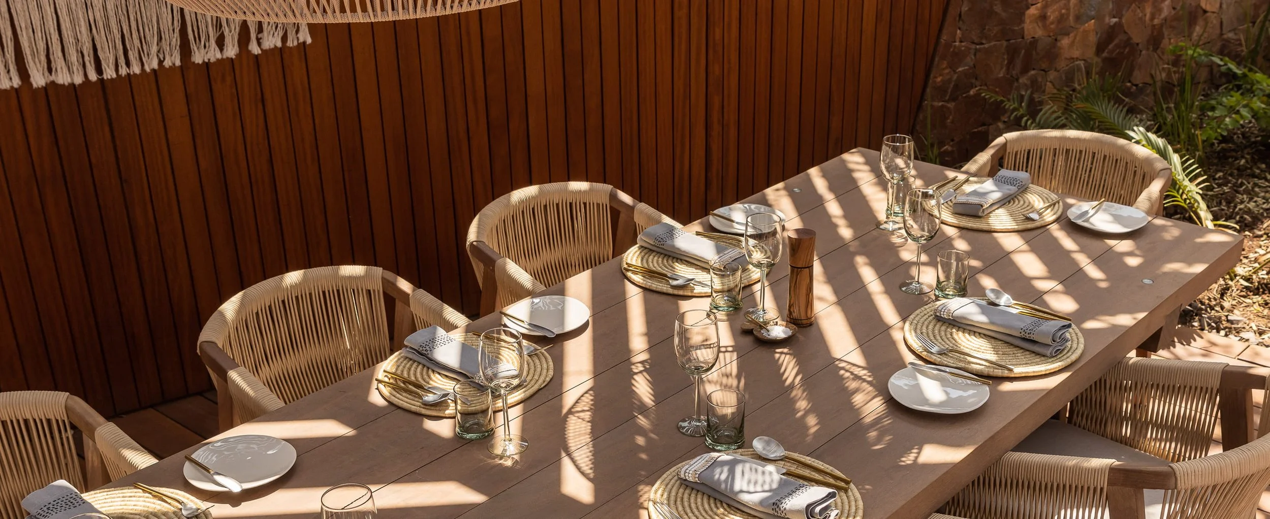An outdoor dining table set for a meal with plates, napkins, silverware, wine glasses, and water glasses, in a shaded area with a wooden wall and plants nearby.
