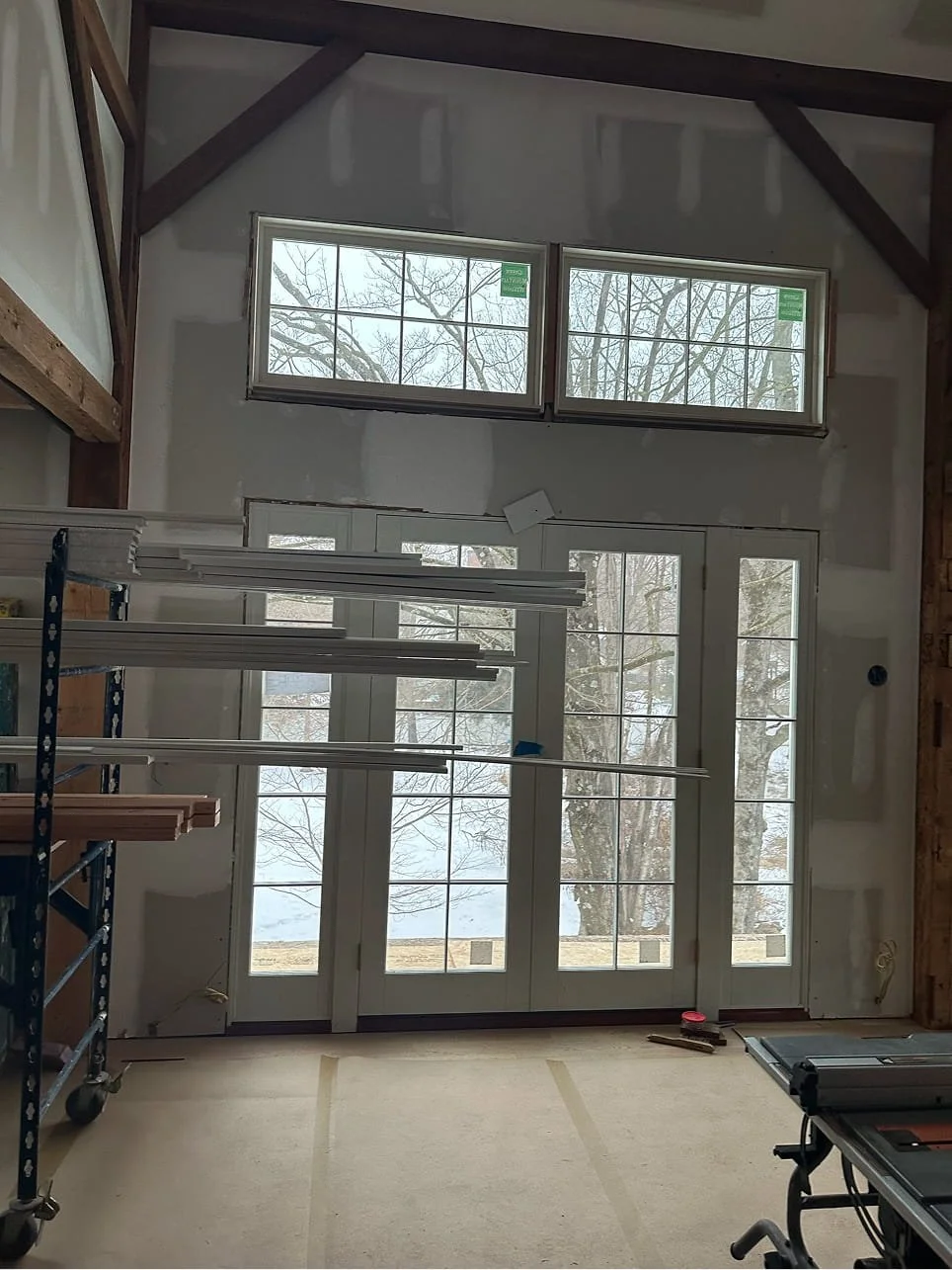 Interior of a room under construction with large windows and French doors facing outside, showing a snowy landscape with trees.
