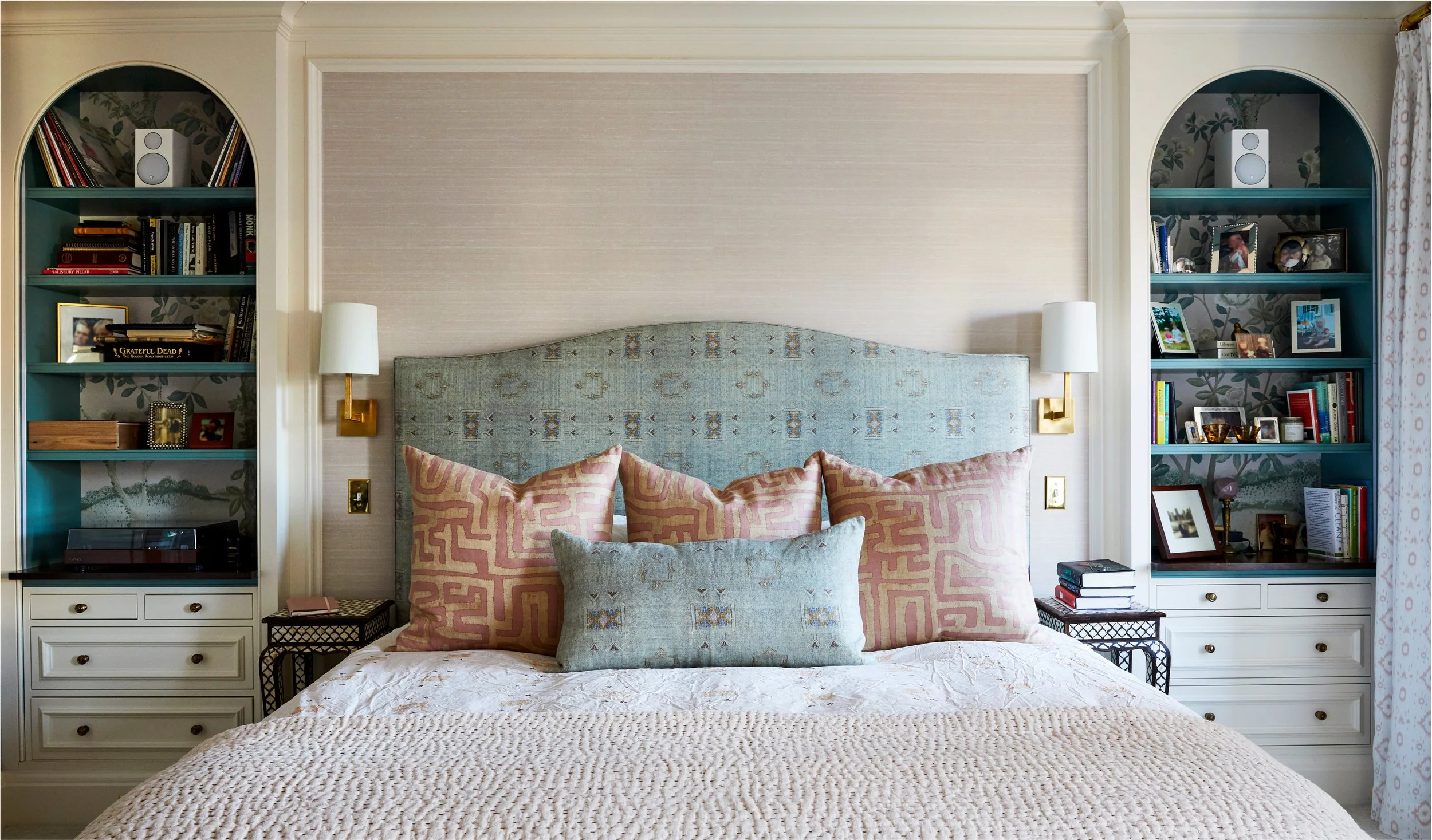 A bedroom with a bed, decorative pillows, built-in bookshelves on each side, and framed photos. The headboard is upholstered and a small table with books is at the foot of the bed.