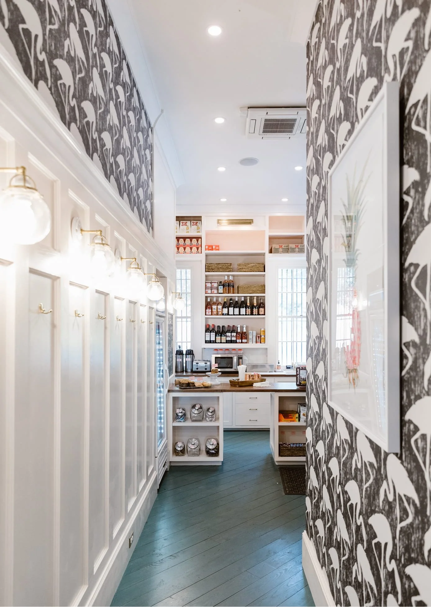 Interior view of a cozy café or small restaurant with white cabinets, stocked shelves of bottles, and patterned wallpaper featuring flamingos, illuminated by ceiling lights.