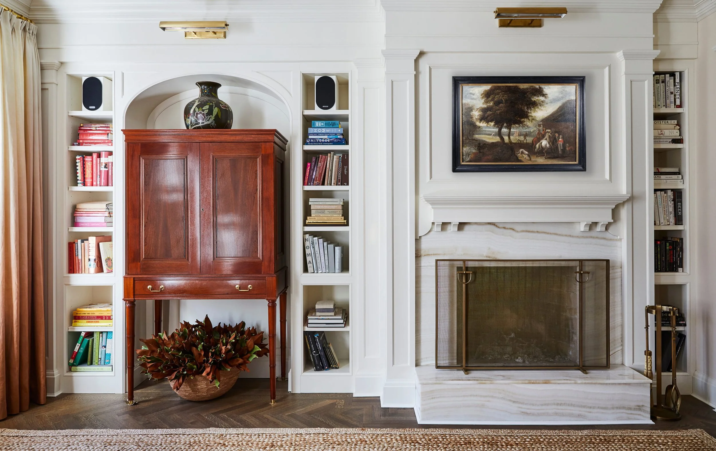 A living room with a white built-in bookshelf surrounding a fireplace, decorated with a framed landscape painting, a large black and floral ceramic vase on a red wooden cabinet, a plant in a basket, and golden wall sconces.