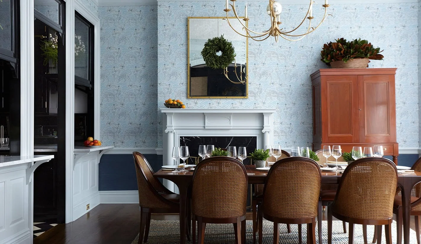 A dining room with a wooden dining table set with multiple wine glasses, potted plants, and a fruit bowl. The room features a white fireplace, a large mirror, a chandelier, and a wooden cabinet with a potted plant on top, all against patterned blue wallpaper.