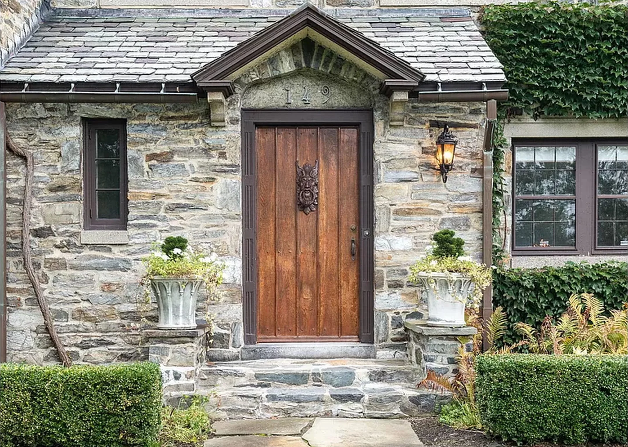 Front view of a stone house with a wooden door, two small windows on each side, and a gabled roof. There are decorative planters with plants on each side of the front steps, a lantern-style light fixture on the right side of the door, and greenery in the background.
